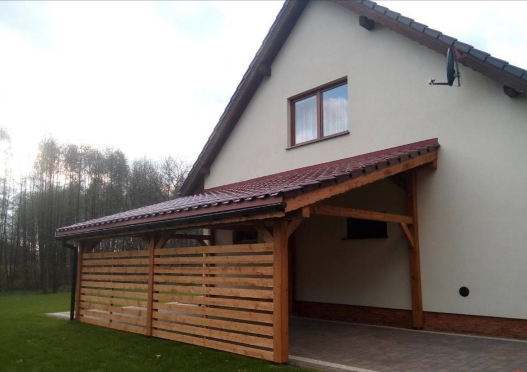 Drewniana wiata garażowa z płotem lamelowym, przylegająca do otynkowanego domu z brązową dachówką, widoczna antena satelitarna.