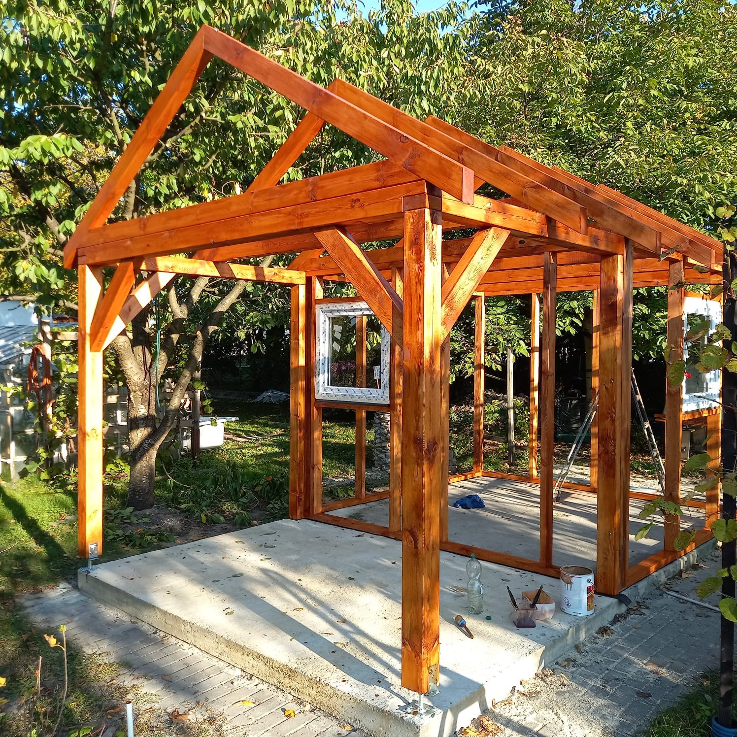 Drewniana konstrukcja altany ogrodowej w trakcie budowy, świeżo pomalowana na brązowo, widoczne narzędzia malarskie i puszka farby na betonowej podstawie, w tle zieleń drzew.
