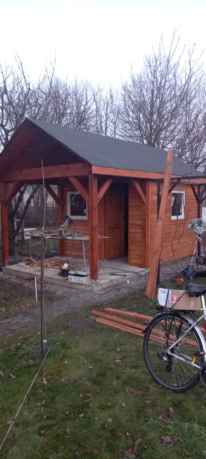 Wznoszona drewniana altana z czarnym dachem, z widocznymi narzędziami pracy, rowerem oraz elementami konstrukcyjnymi na trawniku w pochmurny dzień.
