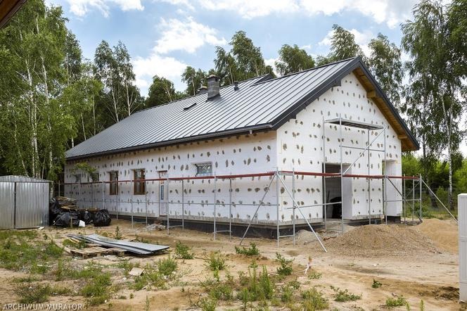 Dom jednorodzinny w trakcie docieplania ścian zewnętrznych styropianem, widoczne rusztowania, fragmenty niezabezpieczonej elewacji i metalowe poszycie dachu na tle drzew