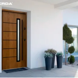 Nowoczesne drzwi zewnętrzne z drewna, z przeszkleniem pionowym i poziomymi frezowaniami, marki Gerda, zamontowane w białym budynku z roślinami w donicach.