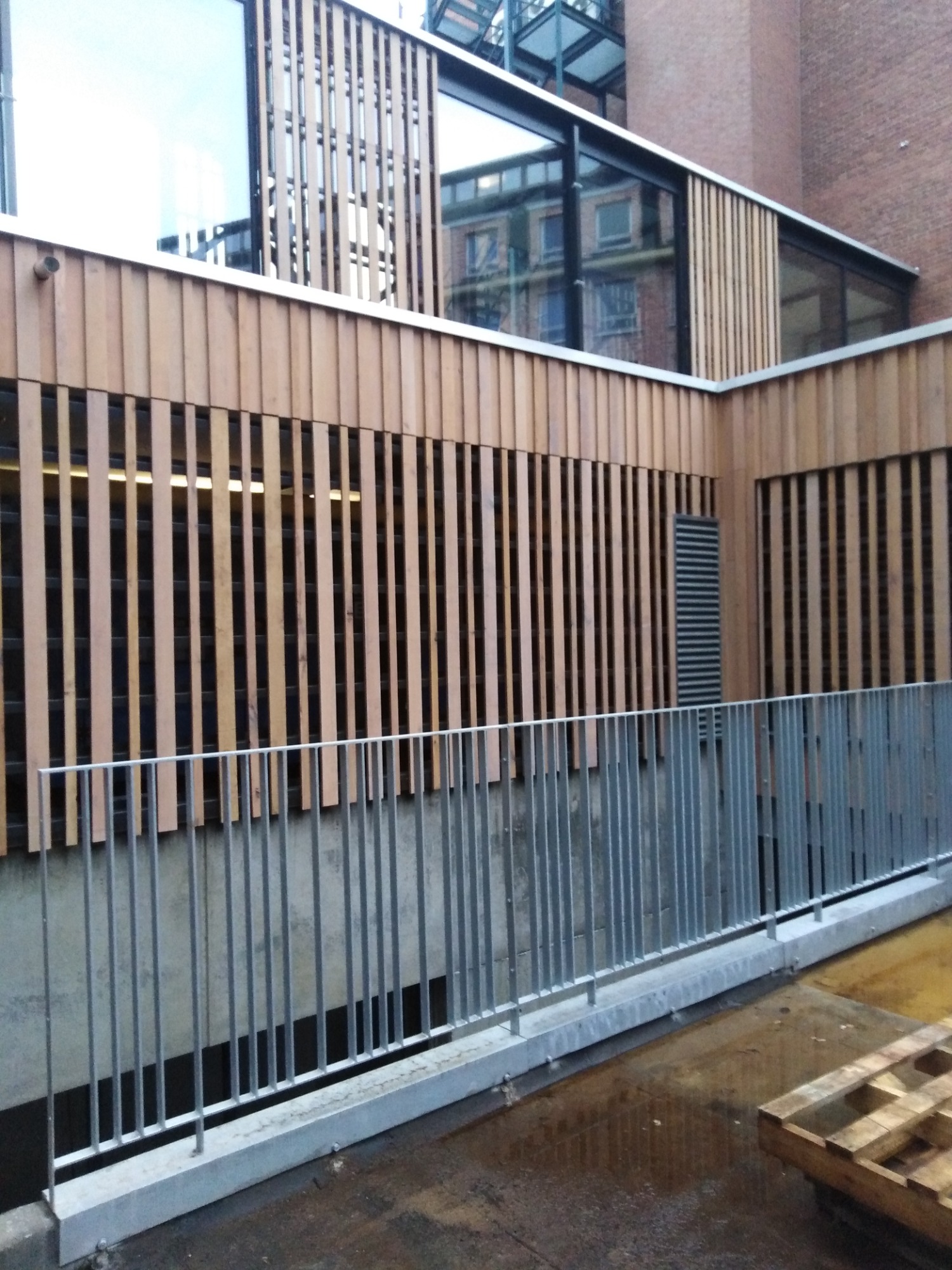 Fasada budynku z drewnianymi panelami pionowymi i metalową balustradą, widok z dołu na fragment budynku z cegły w tle.