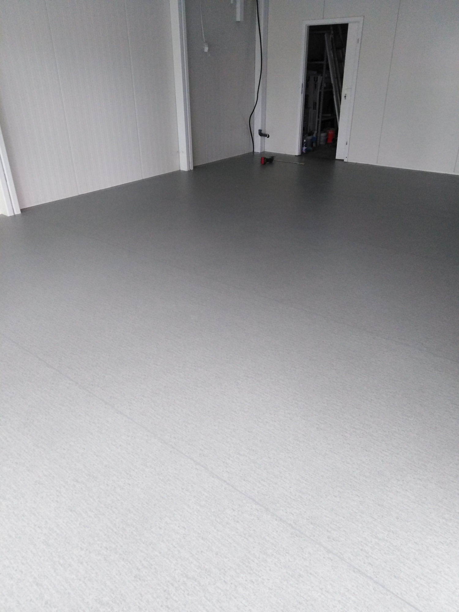 Jasnoszara podłoga z PCV w nowo wykończonym pomieszczeniu z białymi panelami ściennymi i otwartymi drzwiami, przez które widać drabinę i narzędzia.