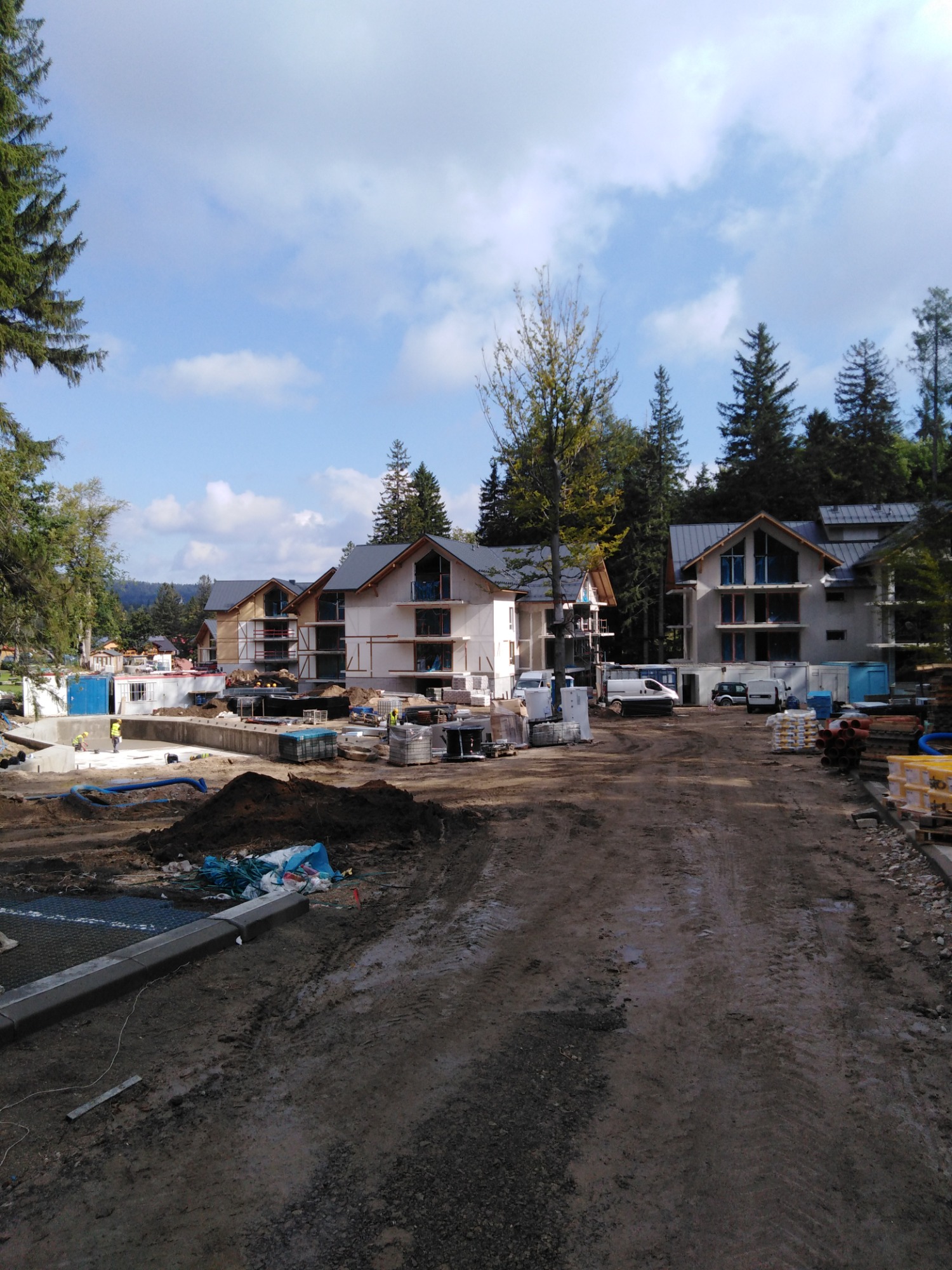 Wczesny etap budowy osiedla domów murowanych z widocznymi rusztowaniami, częściowo otynkowanymi ścianami, oraz przygotowanym terenem z materiałami budowlanymi i pracownikami w oddali.