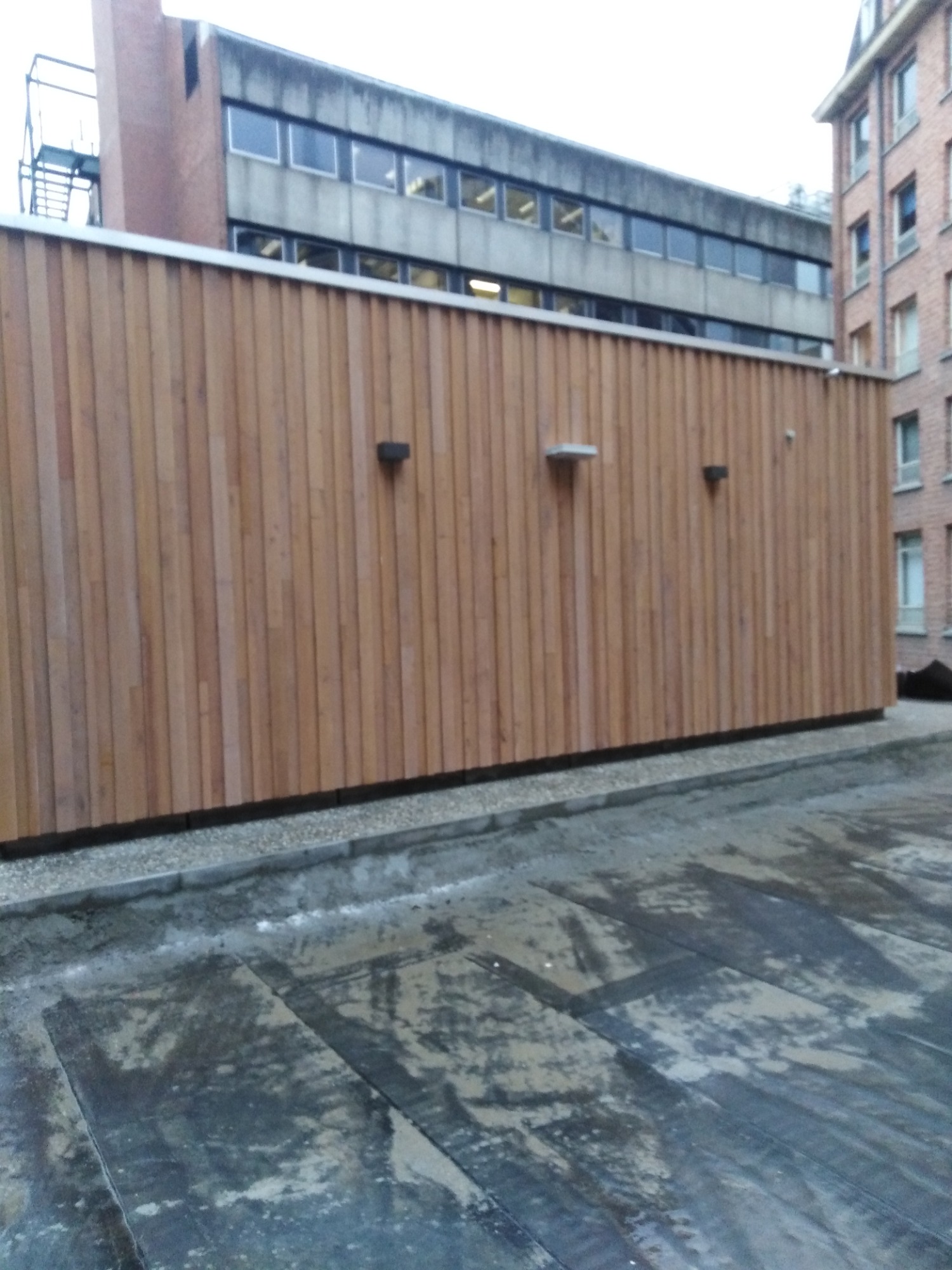 Nowoczesna drewniana elewacja budynku z ukrytymi punktami oświetleniowymi i widoczną konstrukcją dachu oraz budynkami w tle.