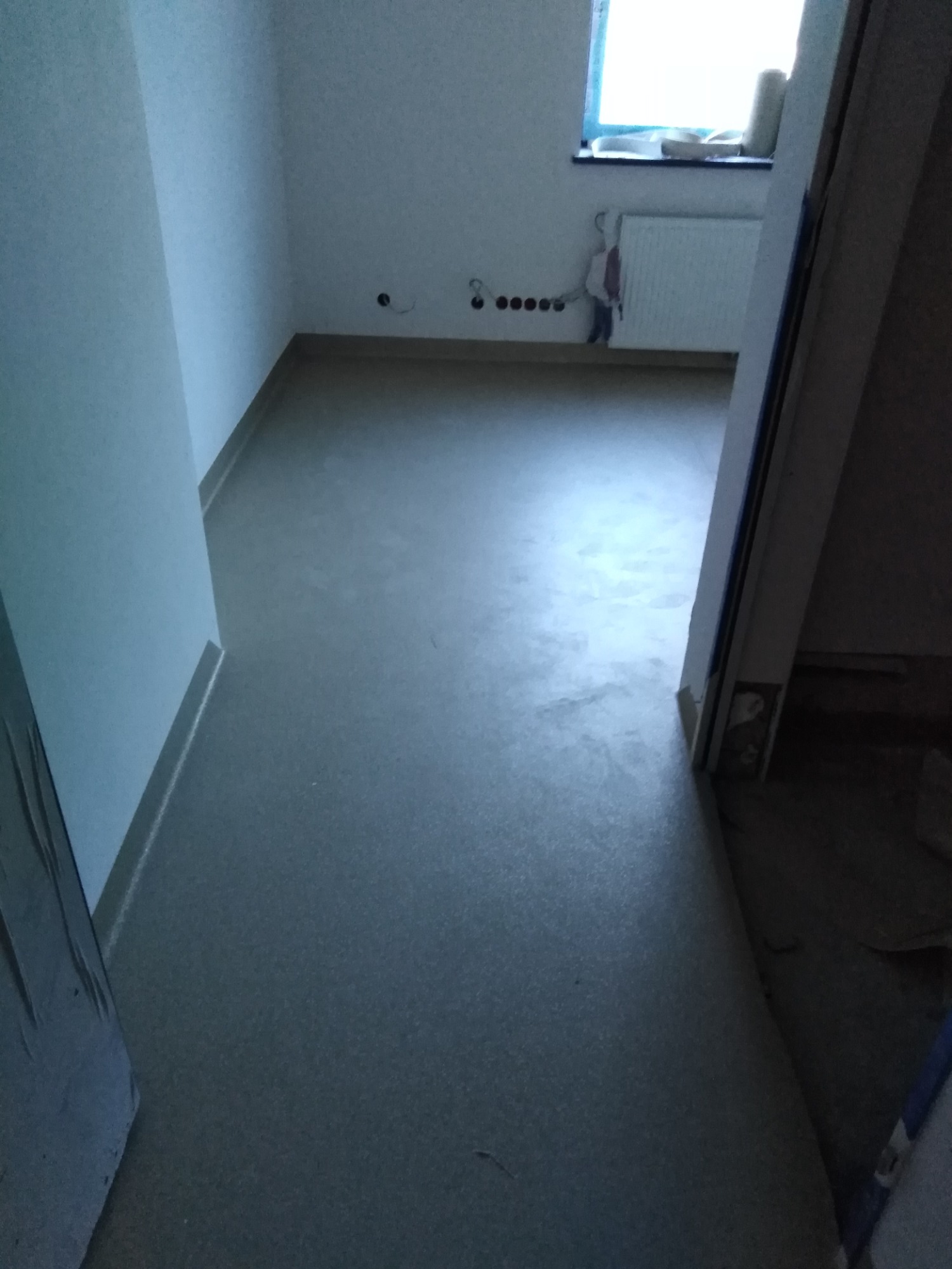 Wnętrze pomieszczenia z nową, jednolitą, szarą wykładziną podłogową; widoczne ślady po instalacji grzejnika, ściany pomalowane na biało.
