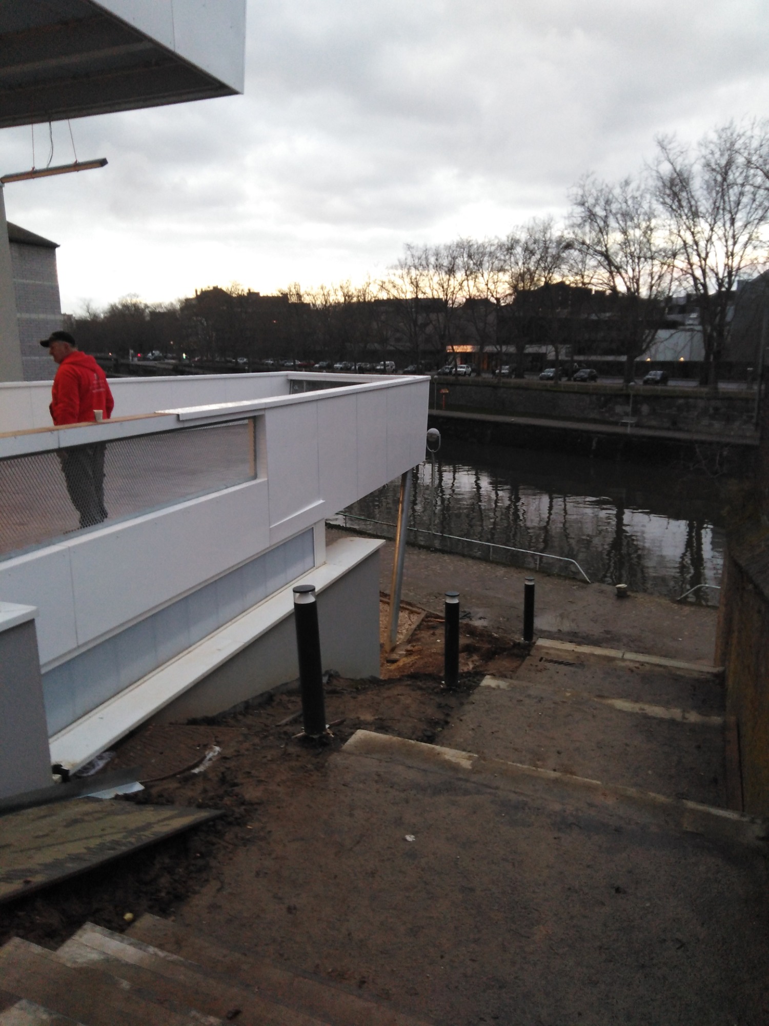 Biała konstrukcja z balustradą nad rzeką, widok z góry na schody i słupki oświetleniowe, mężczyzna w czerwonej bluzie stoi na konstrukcji, pochmurne niebo w tle.