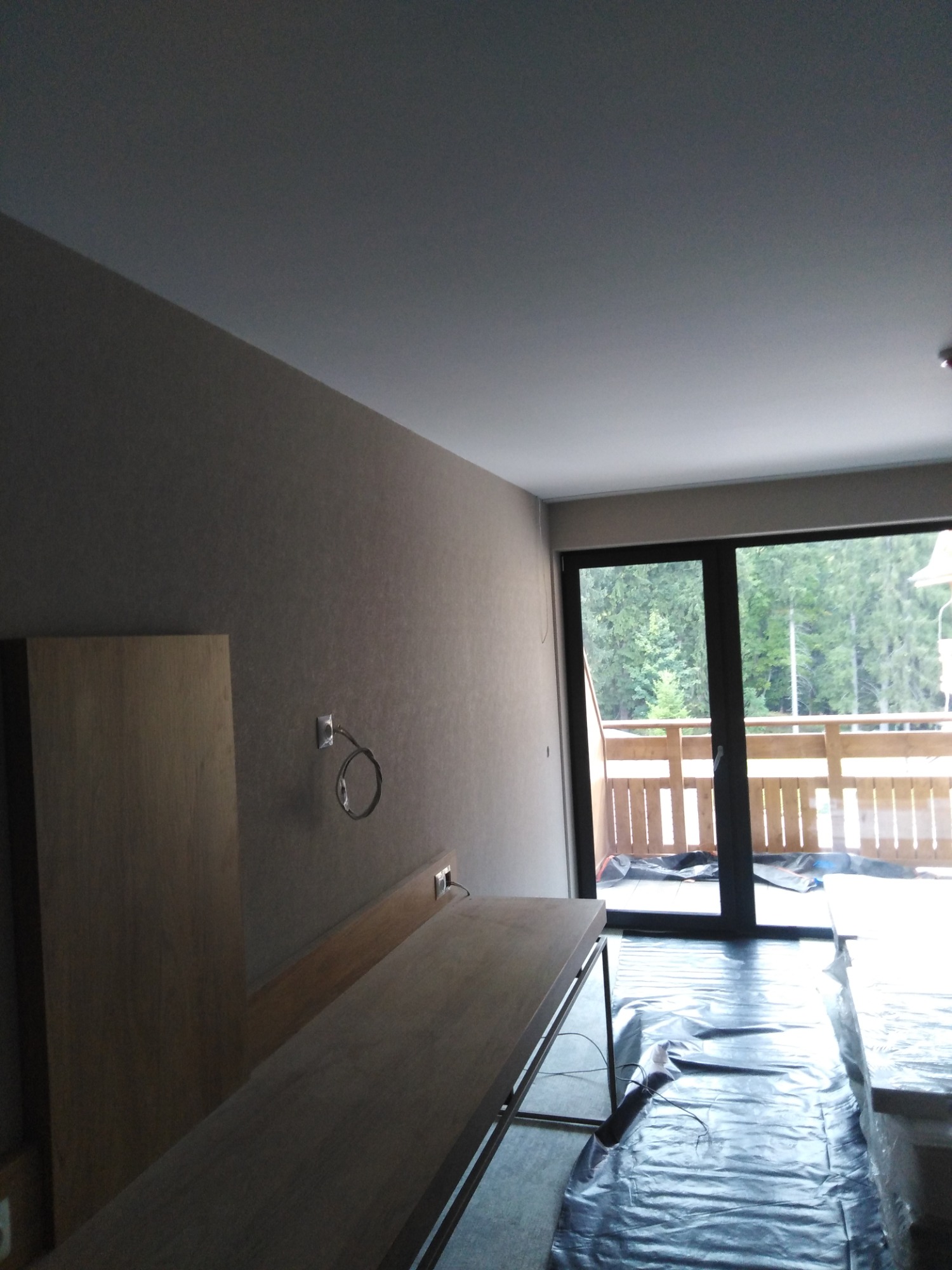 Wnętrze pokoju w trakcie prac wykończeniowych: ściany pomalowane na szaro, podłoga zabezpieczona folią malarską, widoczne gniazdko elektryczne z wystającym przewodem oraz drzwi balkonowe z widokiem...