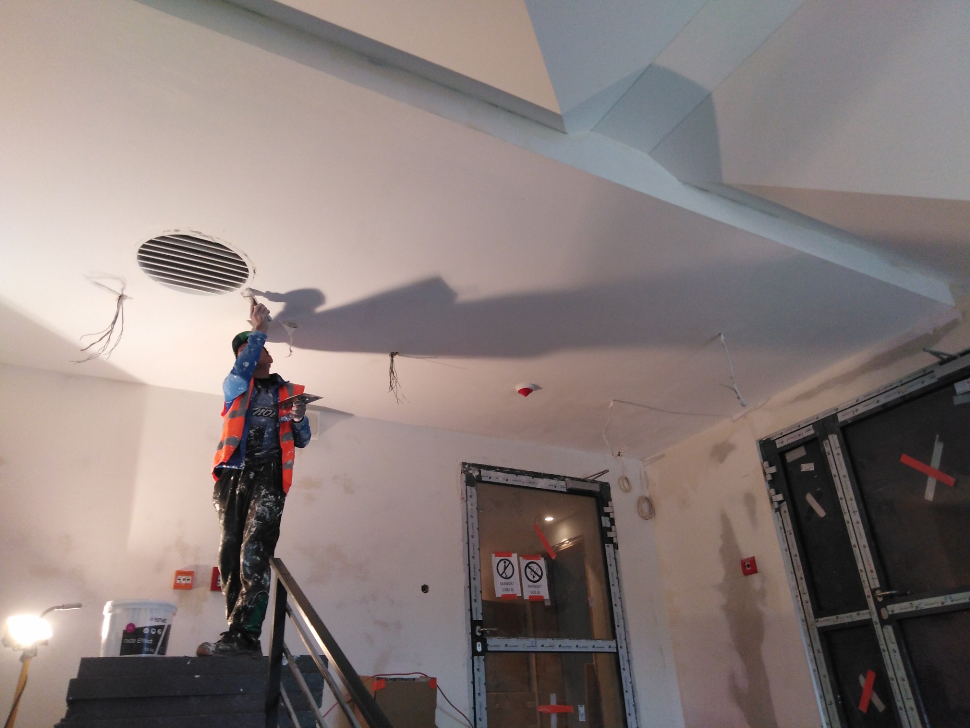 Pracownik w pomarańczowej kamizelce na schodach, szpachluje sufit wokół kratki wentylacyjnej w pomieszczeniu w trakcie remontu. Widoczne nieotynkowane ściany i drzwi z oznaczeniami.