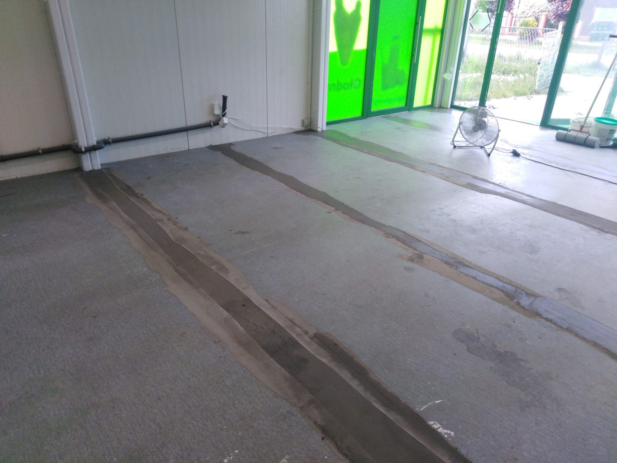 Przygotowanie podłoża pod wykładzinę: szpachlowanie szczelin na betonowej posadzce w pomieszczeniu z zielonymi oknami i wentylatorem.