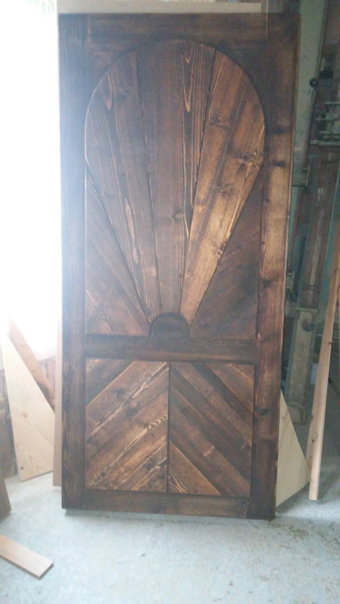 Rustykalne, ręcznie robione drewniane drzwi z motywem wschodzącego słońca i wzorem chevron na dole, w ciemnym odcieniu brązu, stojące w warsztacie.