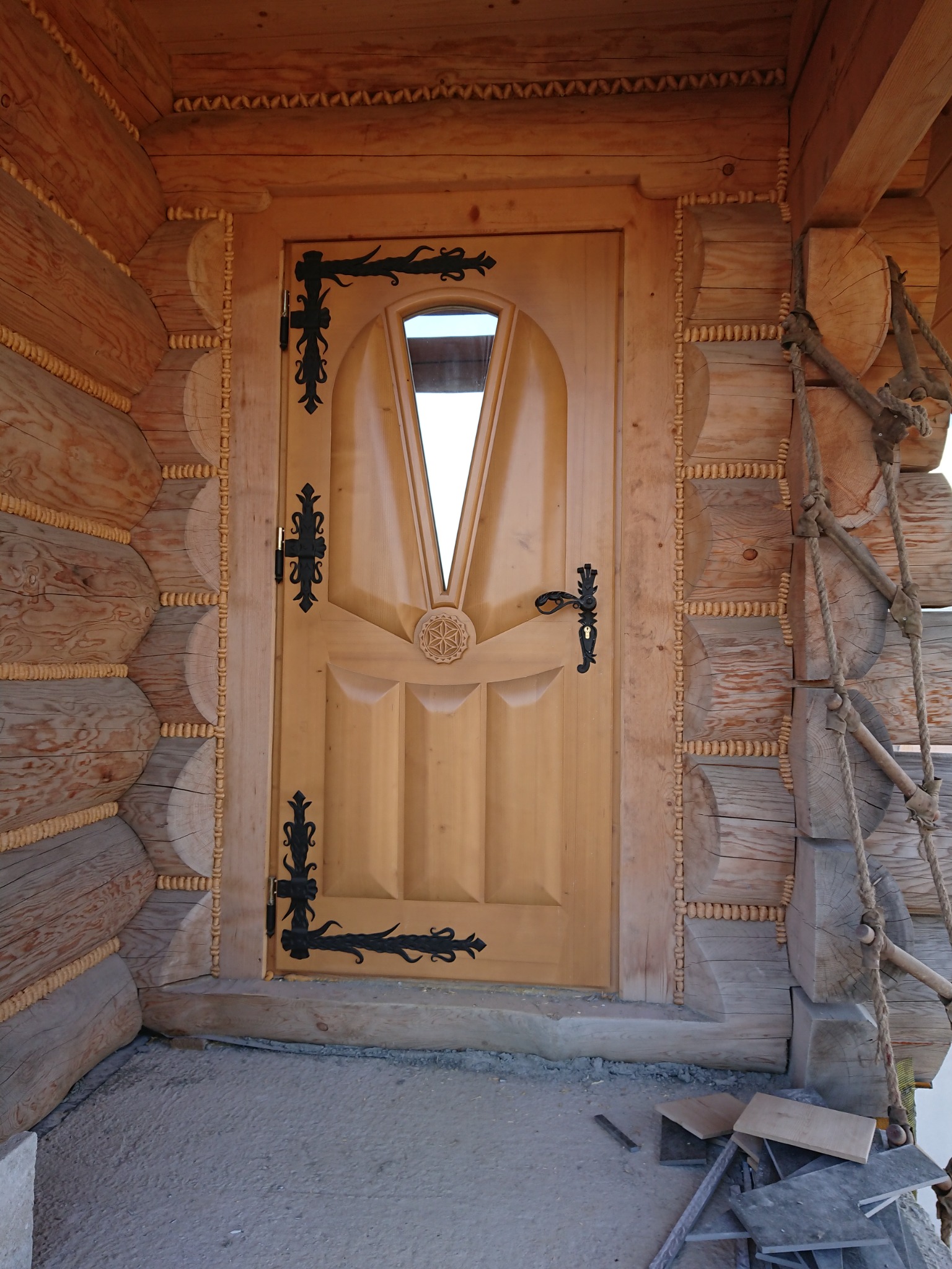 Drewniane wejście do budynku z bali, z ozdobną, jasną, drewnianą stolarką drzwiową i czarnymi, kutymi okuciami. Widoczne detale rzeźbione w drewnie oraz fragment ściany z bali.