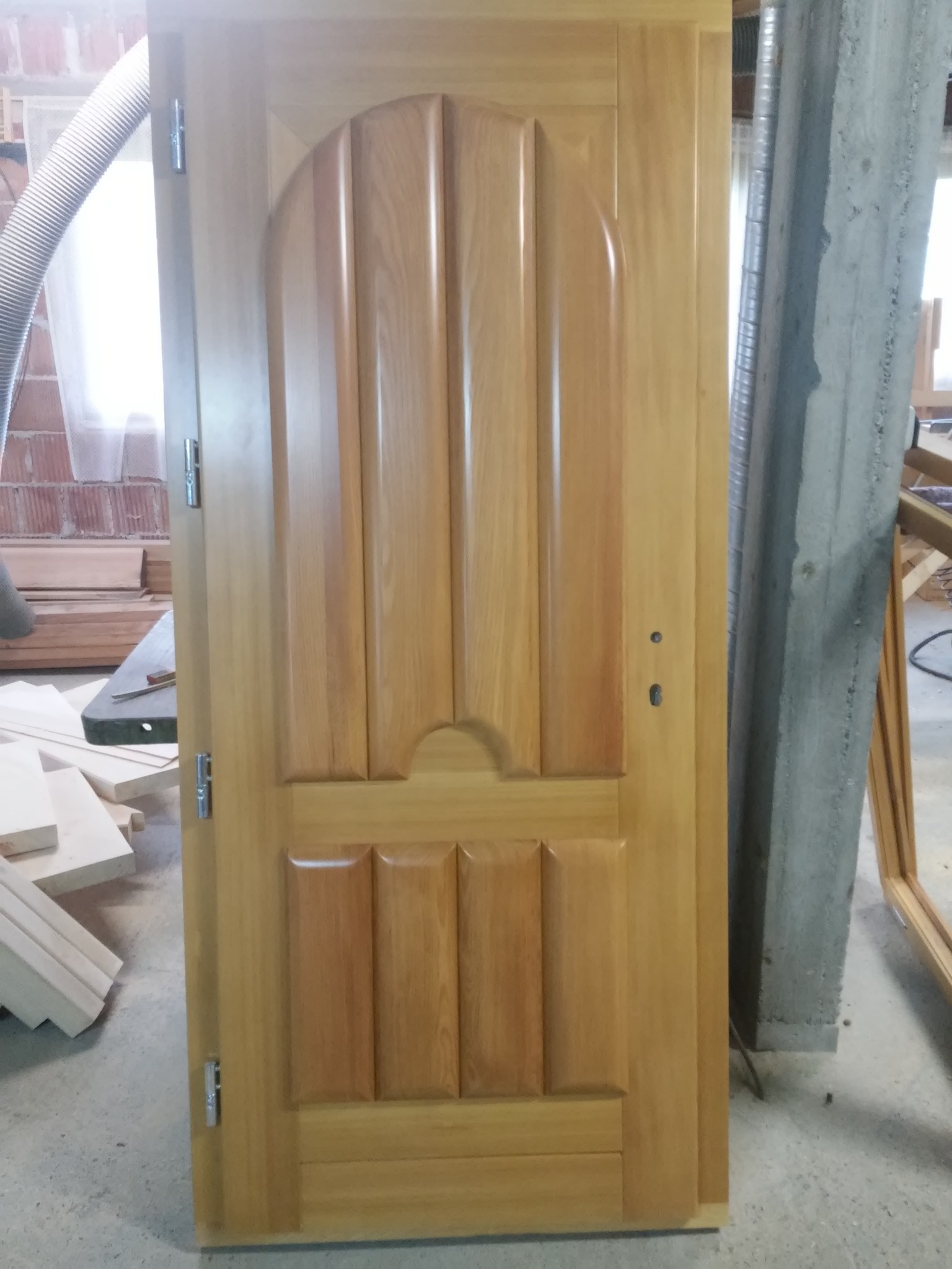 Jasne drewniane drzwi wewnętrzne z dekoracyjnymi panelami, widoczne zawiasy i otwory na zamek, w tle warsztat z elementami stolarskimi i odkurzaczem przemysłowym.