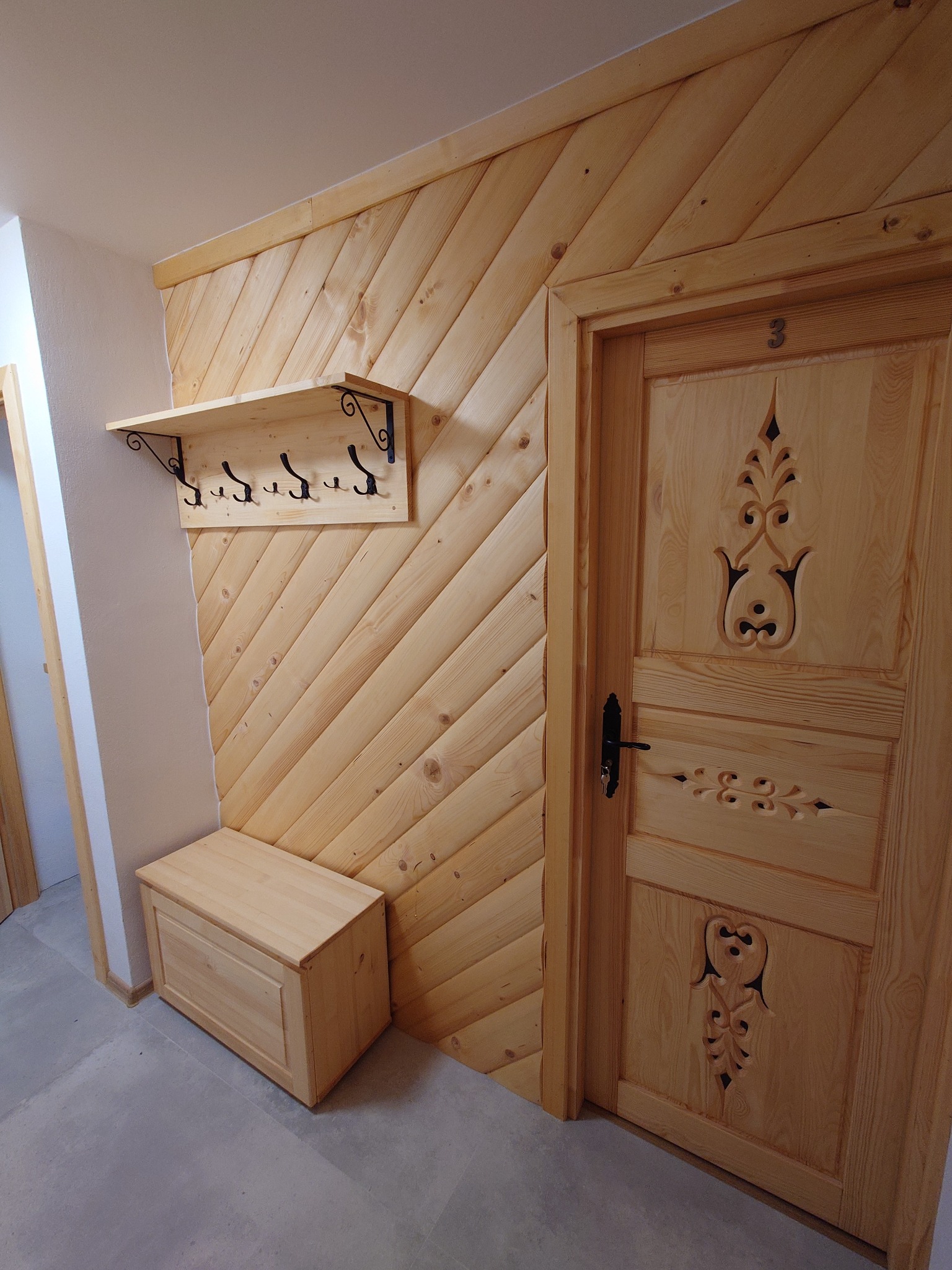 Drewniana ściana wyłożona ukośnie deskami, drewniana półka z wieszakami na czarnych, ozdobnych wspornikach, skrzynia z jasnego drewna i rzeźbione drzwi z numerem '3', tworzące spójny wystrój wnętrza.