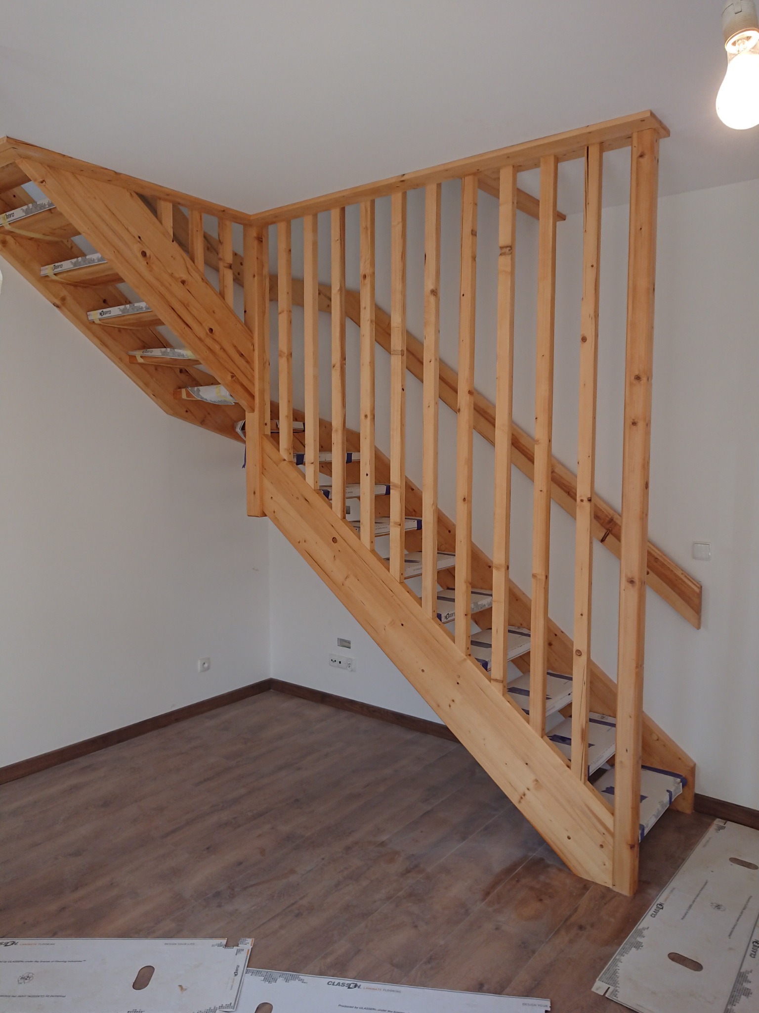 Drewniane schody z balustradą w surowym stanie, zabezpieczone folią na stopniach, widoczne panele podłogowe i fragment ściany z gniazdkami elektrycznymi.