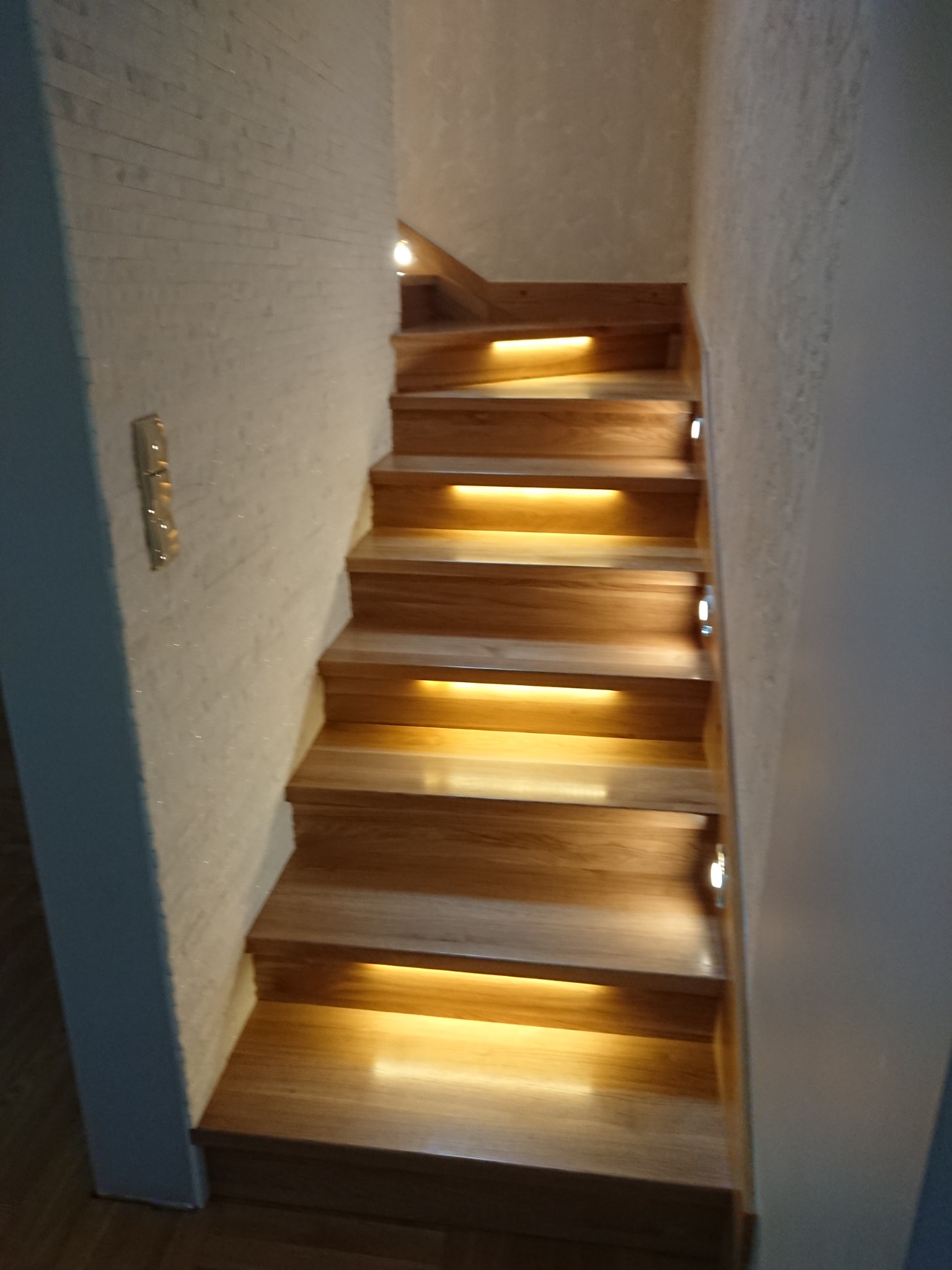Drewniane schody z subtelnym oświetleniem LED wbudowanym w stopnie, ściana wykończona dekoracyjną strukturą, widoczny fragment podłogi.