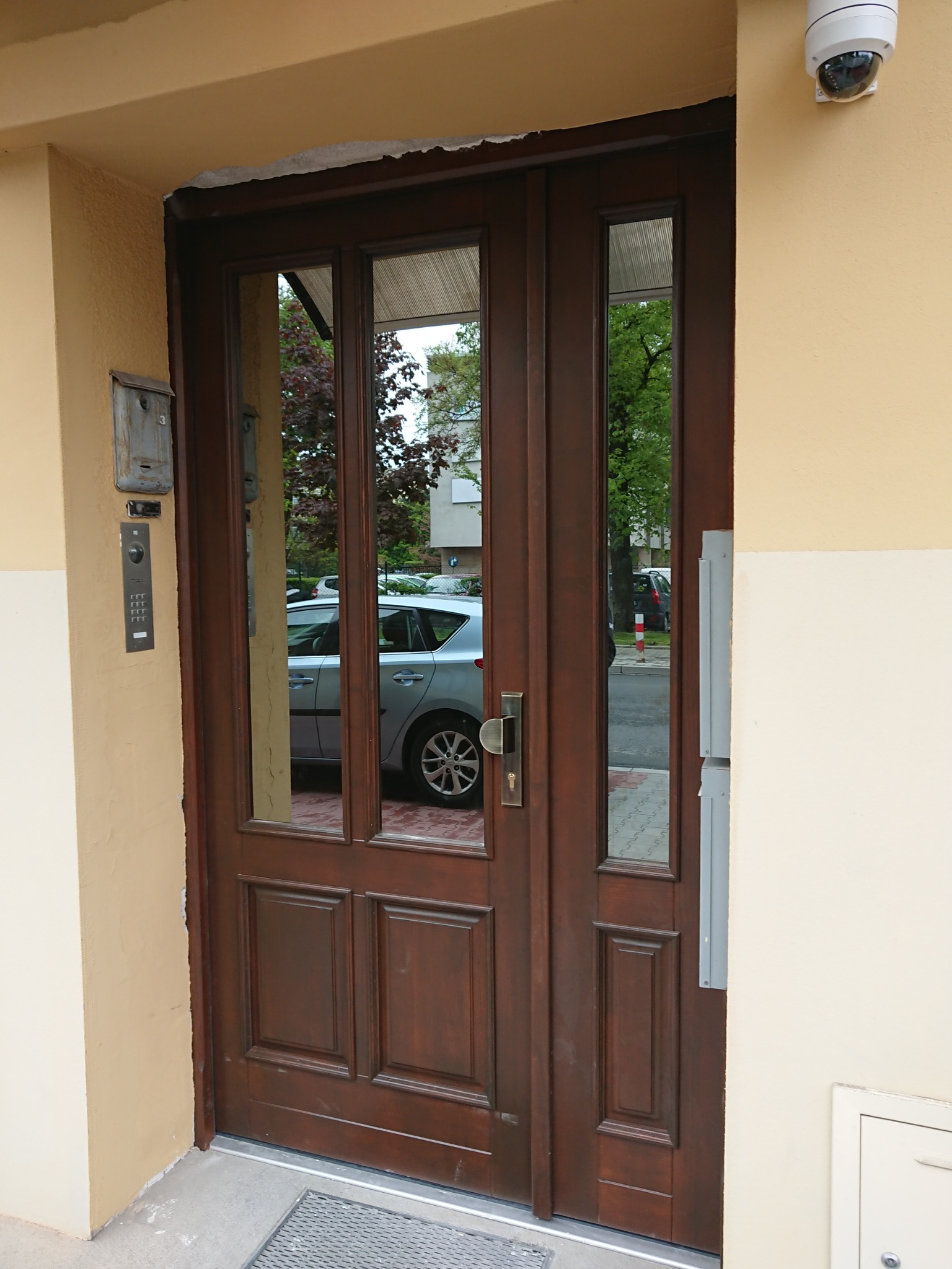 Wejściowe, dwuskrzydłowe drzwi z przeszkleniami w brązowej ramie, zamontowane w budynku z beżową elewacją, widoczna skrzynka na listy i domofon.