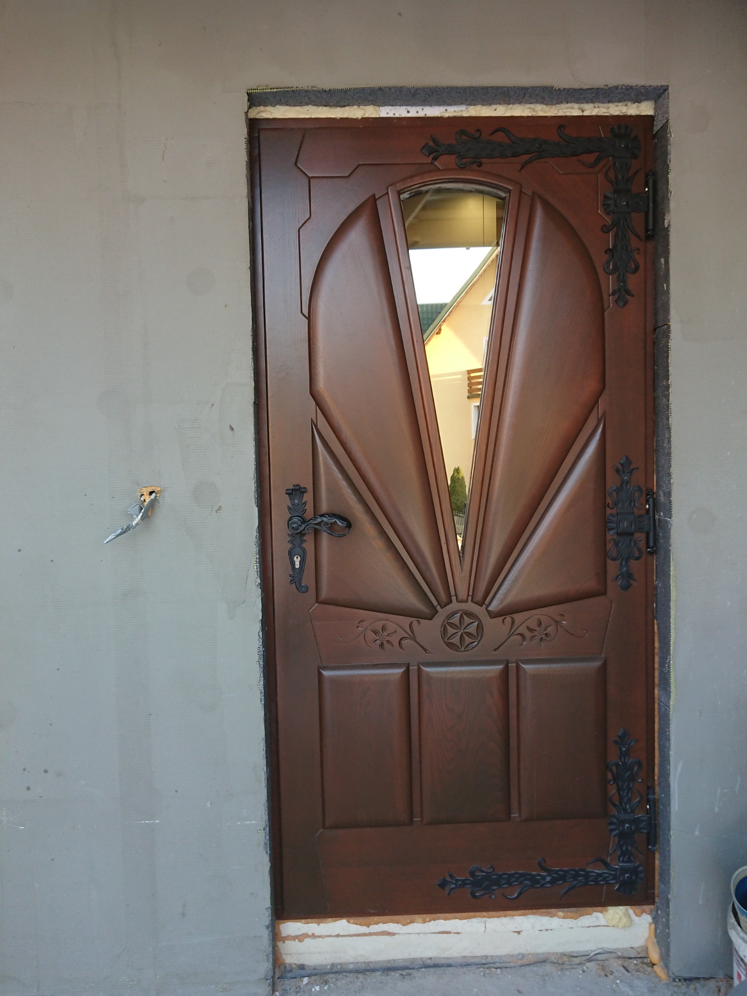 Drewniane drzwi wejściowe z szybą i ozdobnymi metalowymi elementami montowane w surowym otworze ściennym, widoczne narzędzia i niedokończone prace.