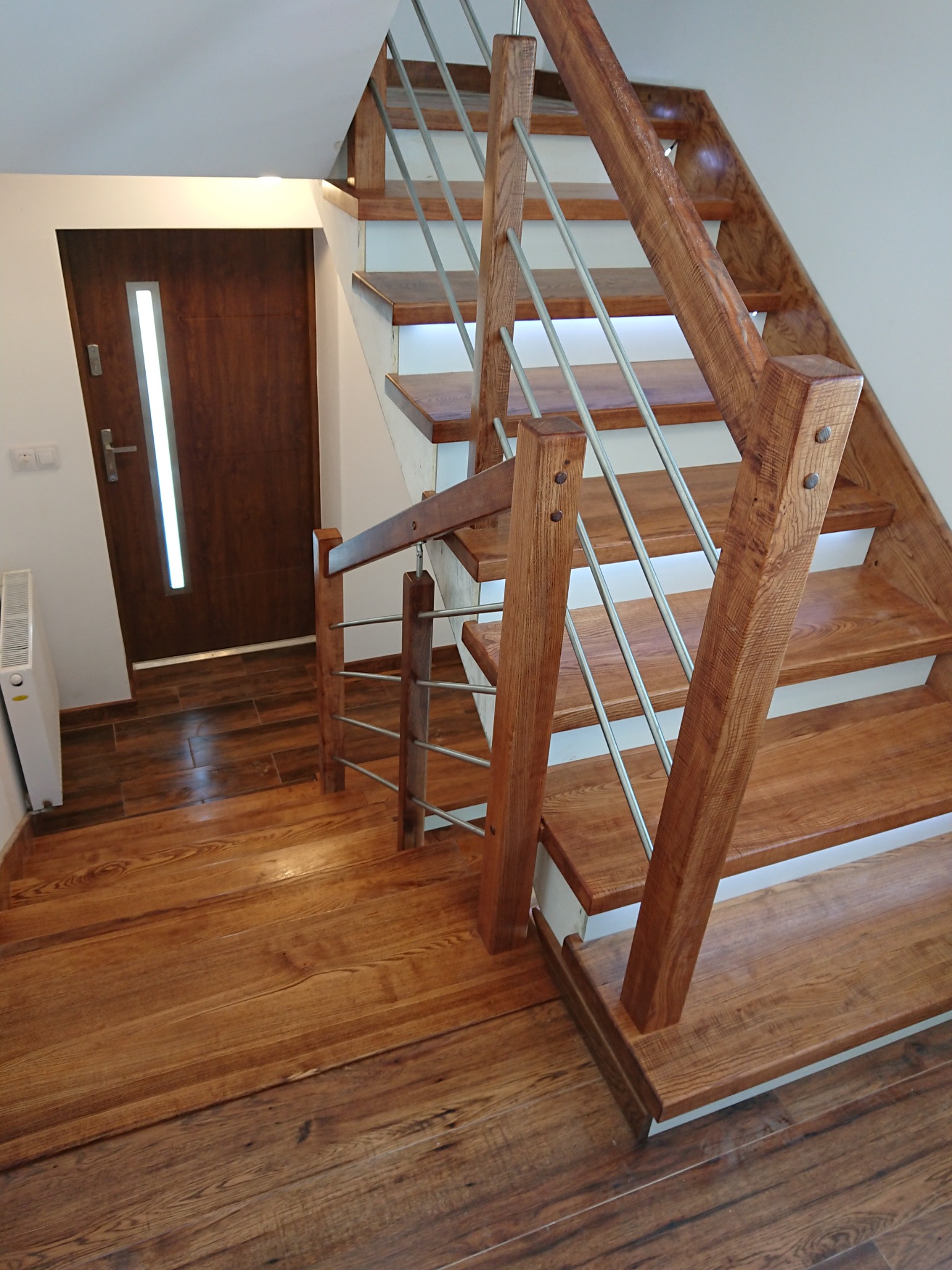 Drewniane schody z metalową balustradą, widok z góry, połączone z podłogą z paneli imitujących drewno, obok drzwi z pionowym przeszkleniem.