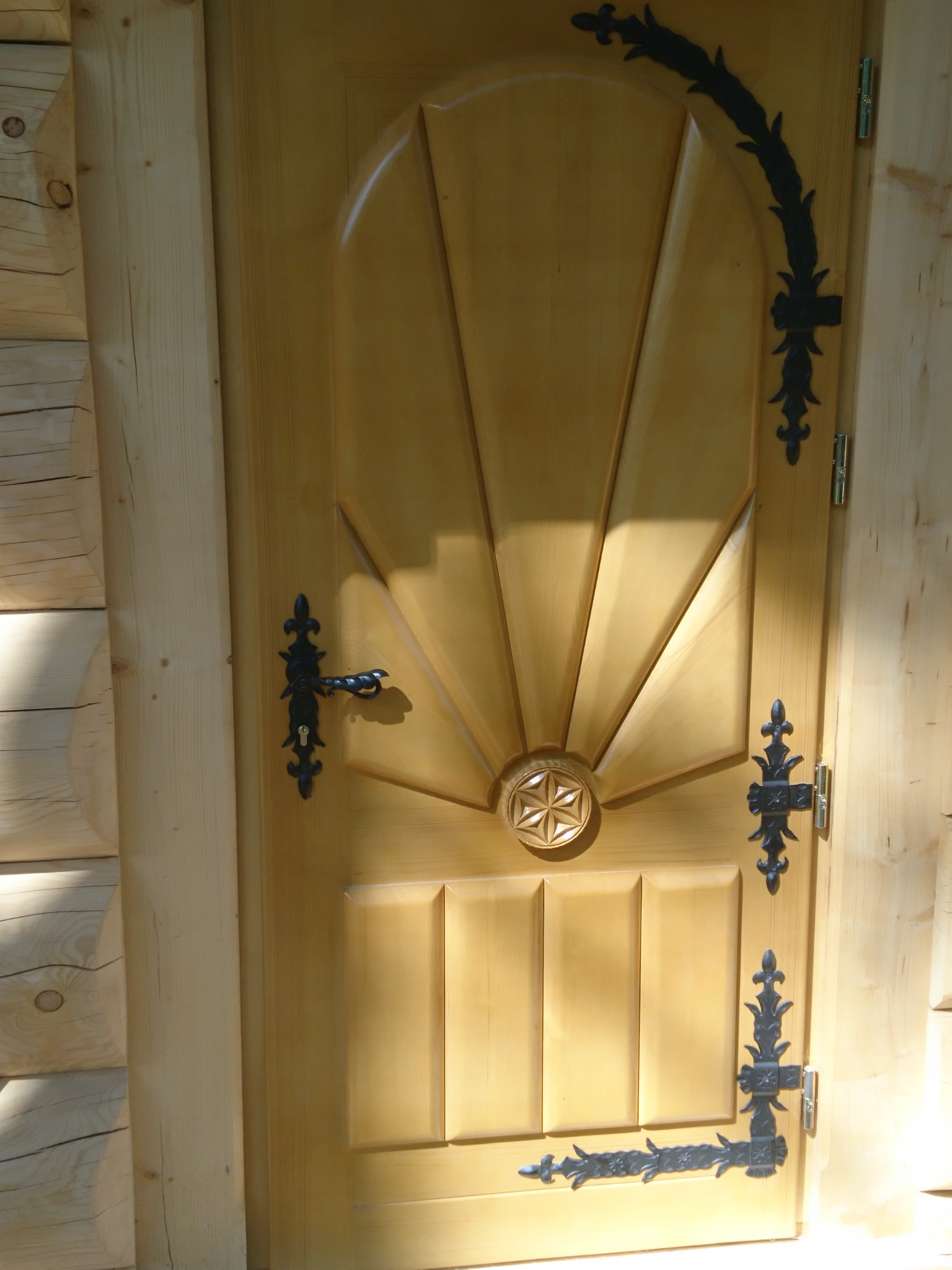 Jasne, drewniane drzwi z dekoracyjnymi, czarnymi okuciami w stylu rustykalnym, zamontowane w drewnianej ścianie z bali.