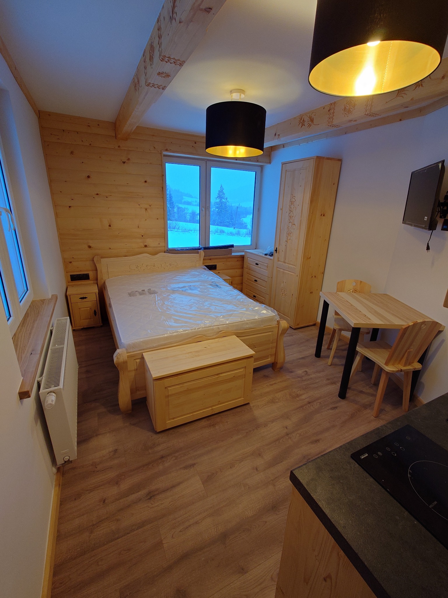 Drewniane wnętrze pokoju z łóżkiem przykrytym folią ochronną, szafą, stolikiem i krzesłami, widok z okna na ośnieżone drzewa.