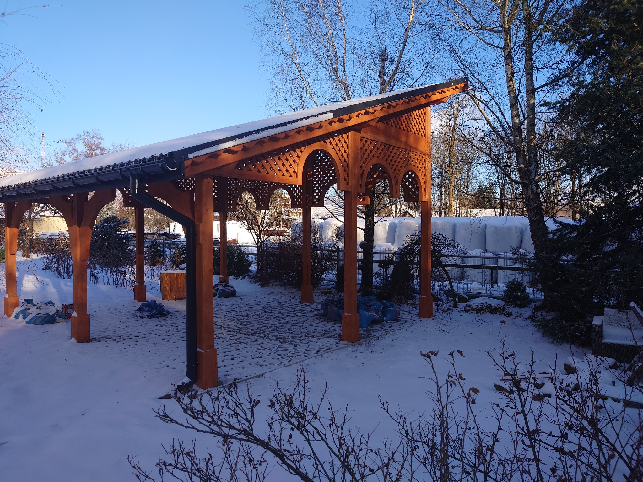 Drewniana konstrukcja z ażurowymi łukami, prawdopodobnie zadaszenie tarasu lub werandy, w zimowej scenerii z pokrytym śniegiem brukowanym podłożem i drzewami w tle.