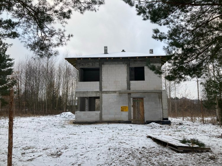 Dwukondygnacyjny dom w stanie surowym otwartym, zbudowany z białych bloczków, na zasypanym śniegiem terenie, z widoczną drewnianą paletą i żółtą tabliczką ostrzegawczą przy drzwiach.