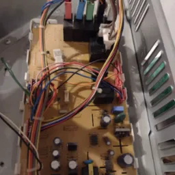 Wnętrze urządzenia AGD z widoczną płytą sterującą, licznymi przewodami w różnych kolorach i elementami elektronicznymi, umieszczoną w metalowej obudowie z otworami wentylacyjnymi.