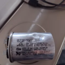 Zbliżenie na aluminiowy kondensator 400VAC 5uF z widocznymi oznaczeniami producenta i parametrami technicznymi leżący na beżowej powierzchni, obok widoczny kabel USB.