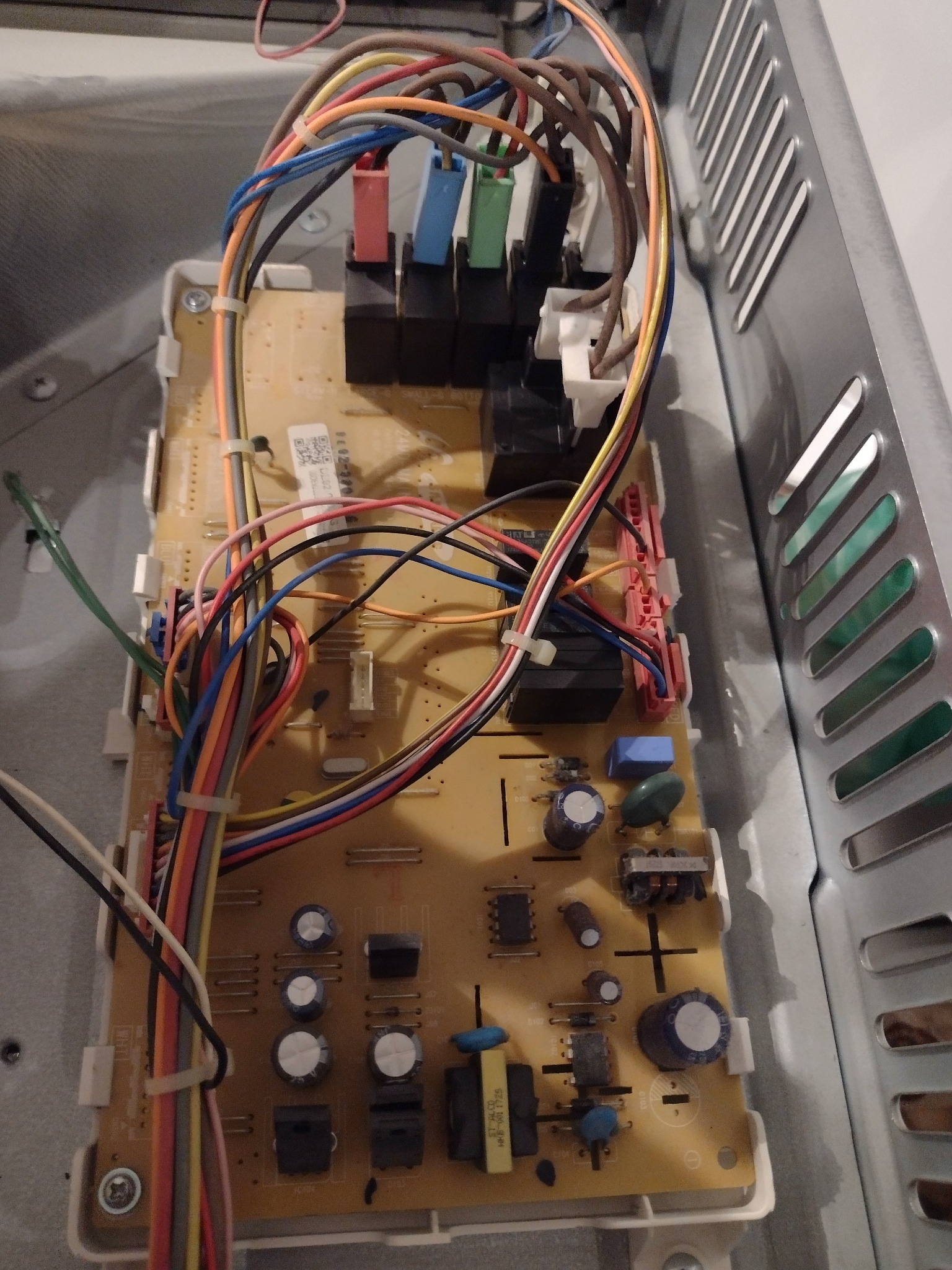 Wnętrze urządzenia AGD z widoczną płytą sterującą, licznymi przewodami w różnych kolorach i elementami elektronicznymi, umieszczoną w metalowej obudowie z otworami wentylacyjnymi.