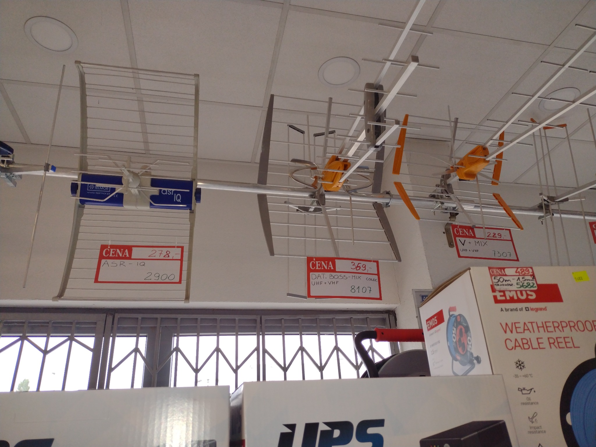 Wystawa anten telewizyjnych w sklepie, widoczne modele ASR-IQ i DAT BOSS-MIX z cenami, na tle sufitu podwieszanego i okna z kratami.