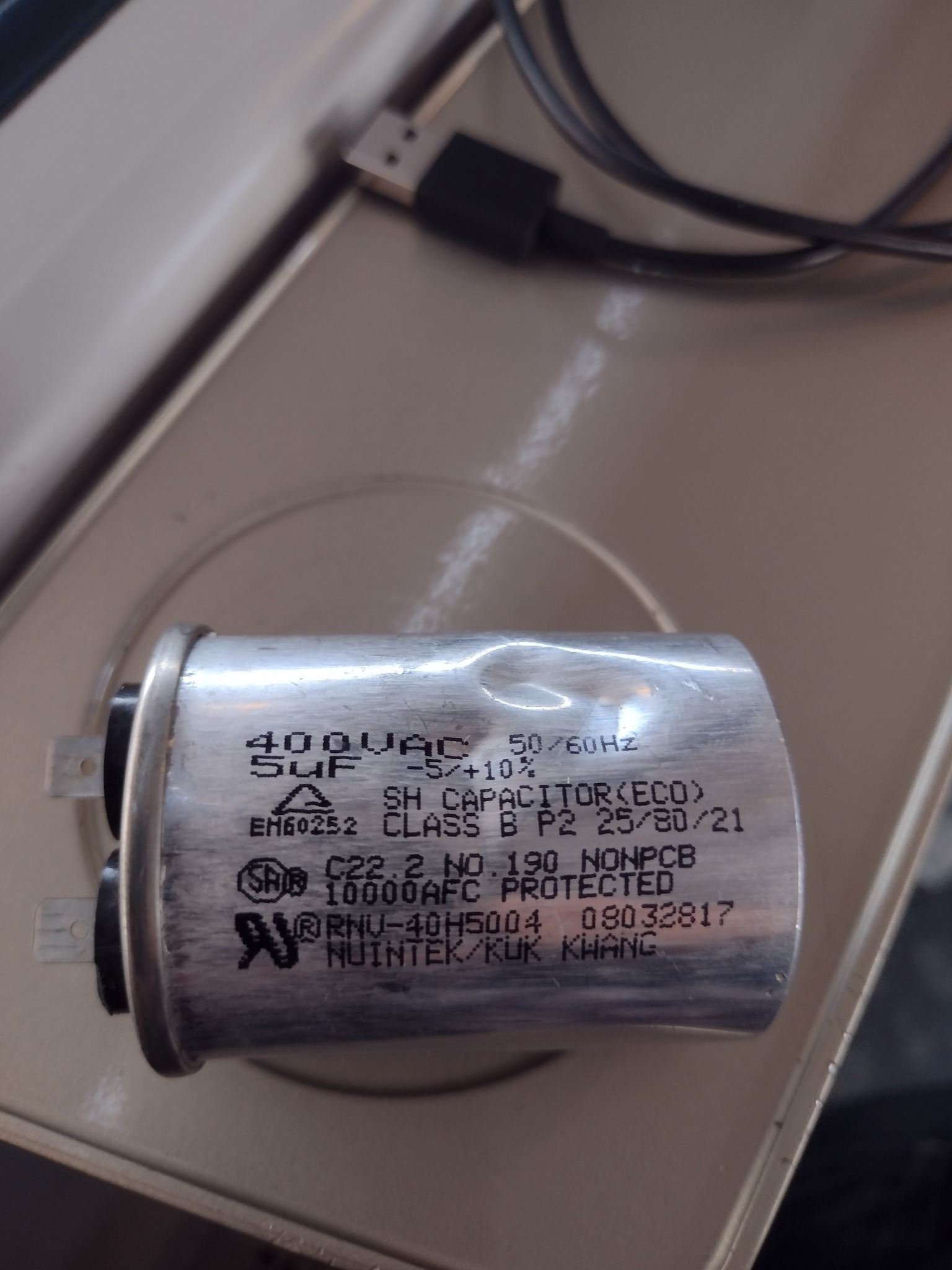 Zbliżenie na aluminiowy kondensator 400VAC 5uF z widocznymi oznaczeniami producenta i parametrami technicznymi leżący na beżowej powierzchni, obok widoczny kabel USB.