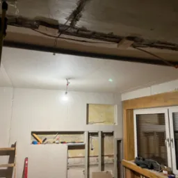 Wnętrze w trakcie remontu: ściany i sufit pokryte płytami, widoczne narzędzia i drabiny, odkryta instalacja elektryczna w suficie, okno z drewnianą ramą.