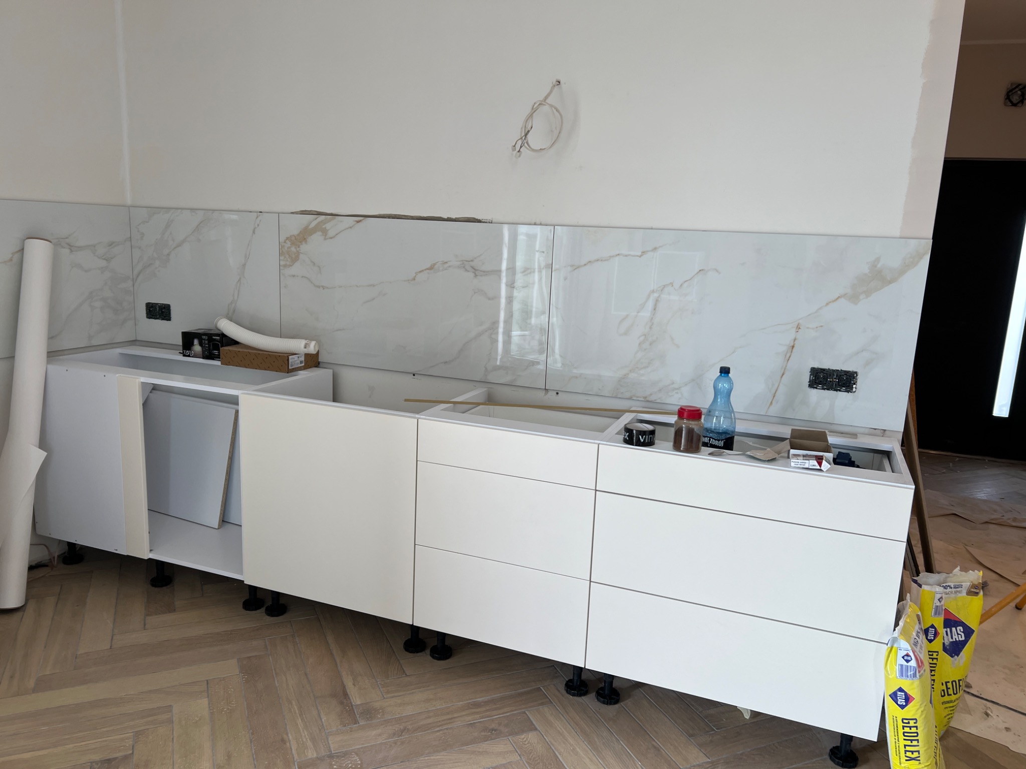 Montaż białych szafek kuchennych z marmurowym wzorem na ścianie, widoczne narzędzia i worki z zaprawą na podłodze z parkietem w jodełkę.