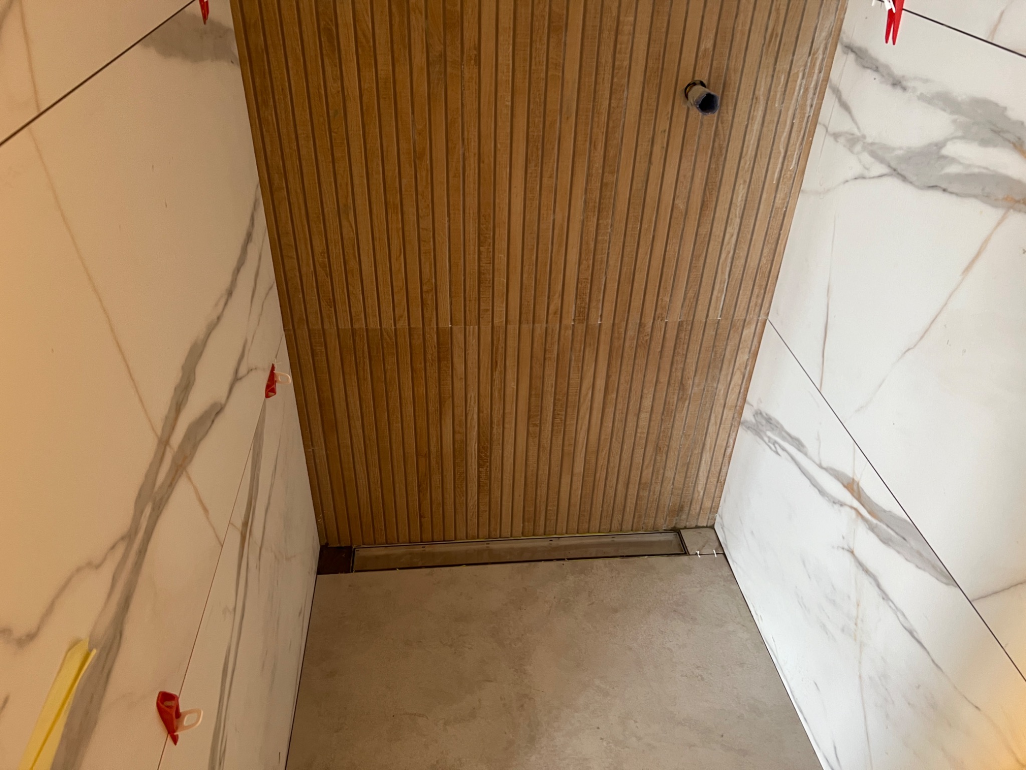 Łazienka w trakcie wykańczania, widoczne płytki imitujące marmur na ścianach, drewnopodobna okładzina z listew na suficie, odpływ liniowy w posadzce, klipsy poziomujące płytki.