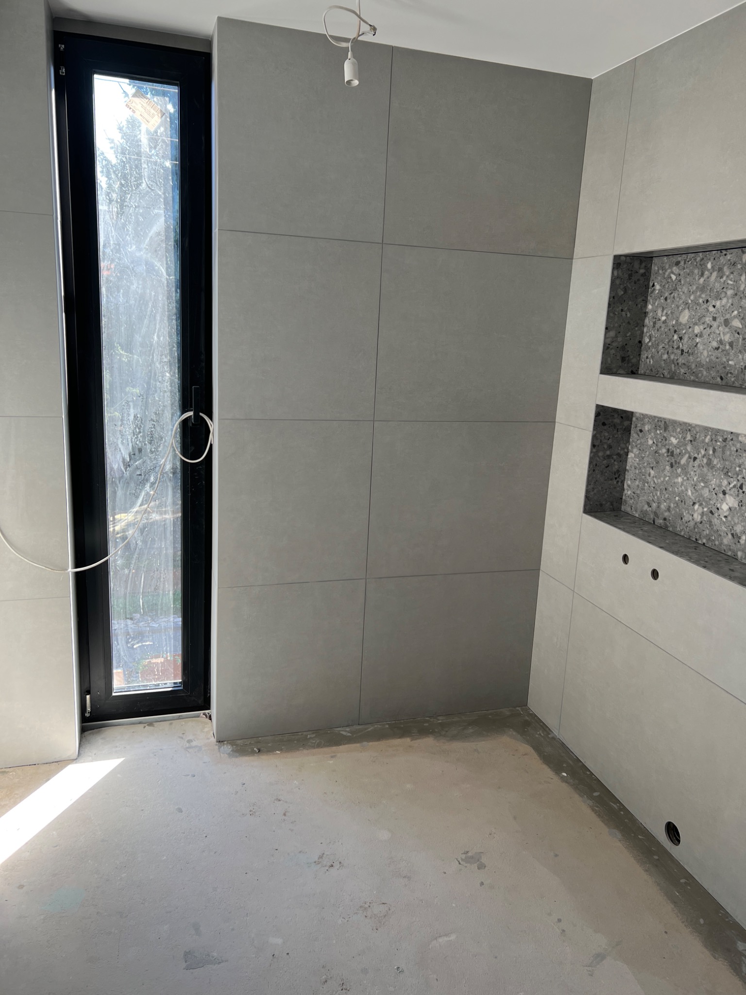 Wnętrze łazienki w trakcie wykańczania, z szarymi płytkami wielkoformatowymi na ścianach, wnękami wykończonymi lastryko i surową betonową podłogą, widoczne okno z czarną ramą.