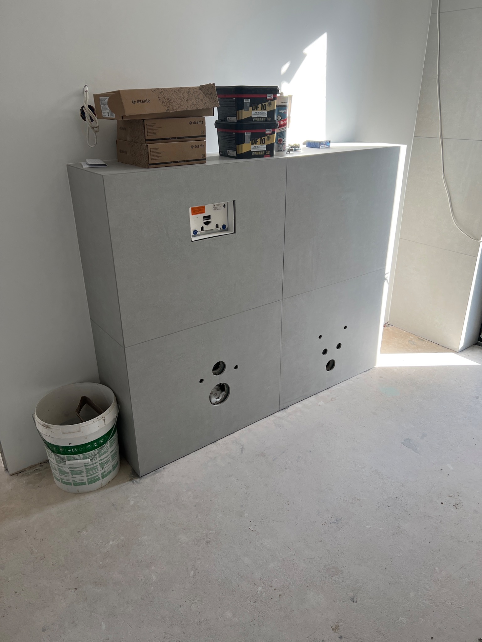 Szara zabudowa WC z płyt w Grodzisku Wielkopolskim, z widocznymi otworami na rury i elementami instalacji, obok stojące opakowania i wiadro, w tle fragment wykończonej ściany.