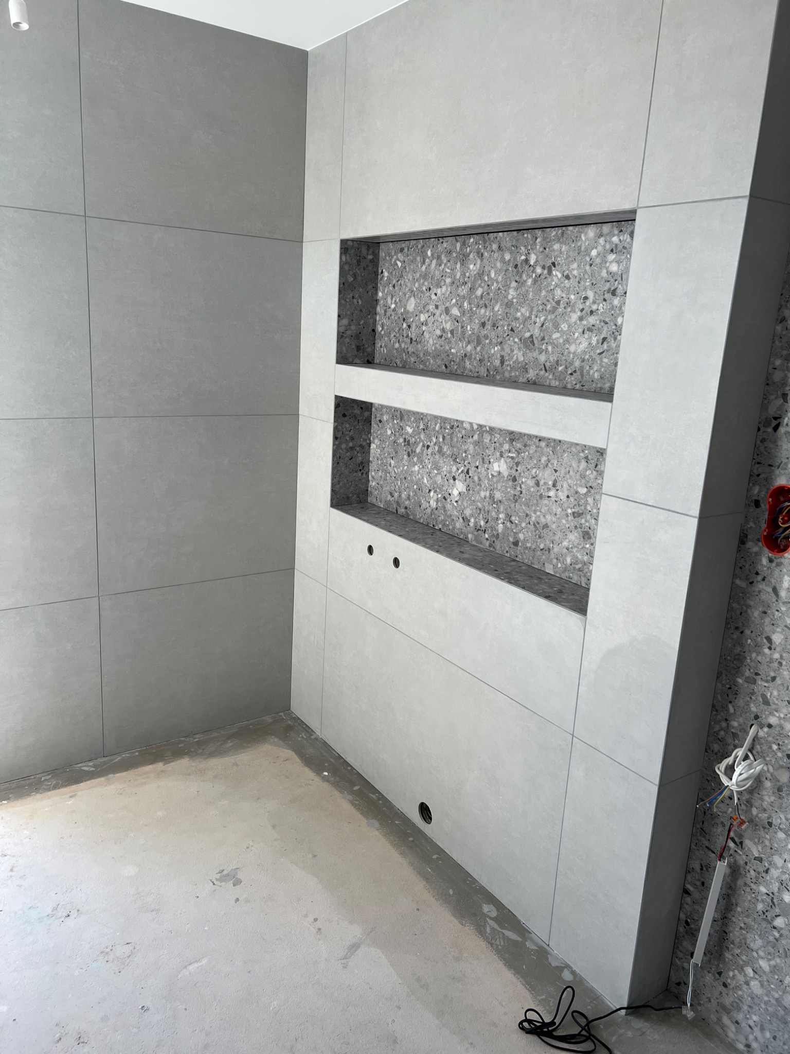 Szara łazienka w trakcie wykończenia, widoczne płytki wielkoformatowe, wnęka z półkami wykończona mozaiką, surowy beton na podłodze, wystające przewody elektryczne.