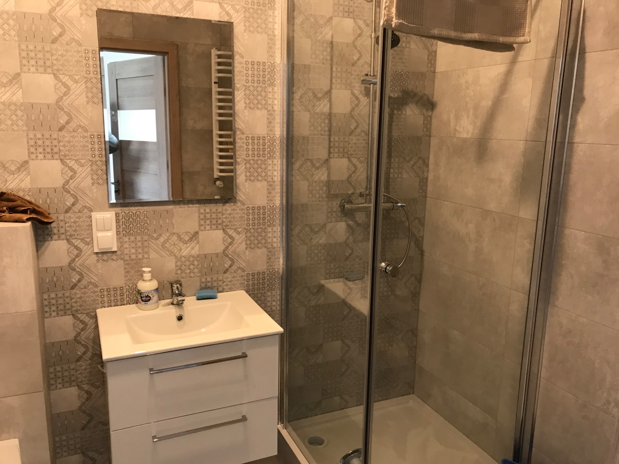 Umywalka z szafką, lustro i kabina prysznicowa w łazience wykończonej płytkami w patchworkowy wzór w odcieniach szarości i beżu.