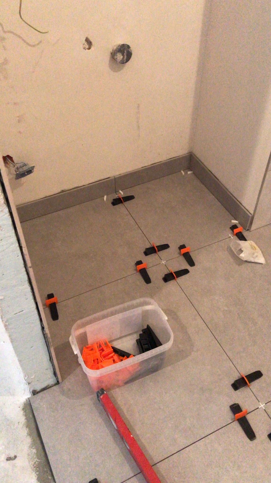 Układanie szarych płytek podłogowych z użyciem poziomicy i systemu poziomowania płytek (klipsy i kliny) w trakcie prac wykończeniowych łazienki, widoczne gniazdka elektryczne w ścianie.