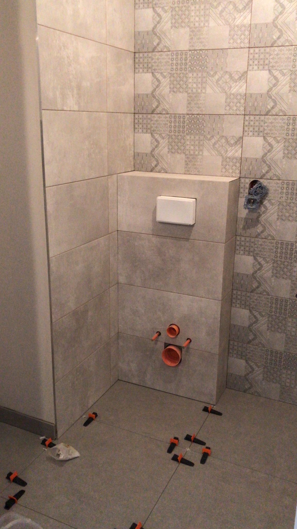 Montaż płytek w łazience, widoczne rury instalacyjne, spłuczka podtynkowa, krzyżyki dystansowe i fragment wzorzystej ściany.