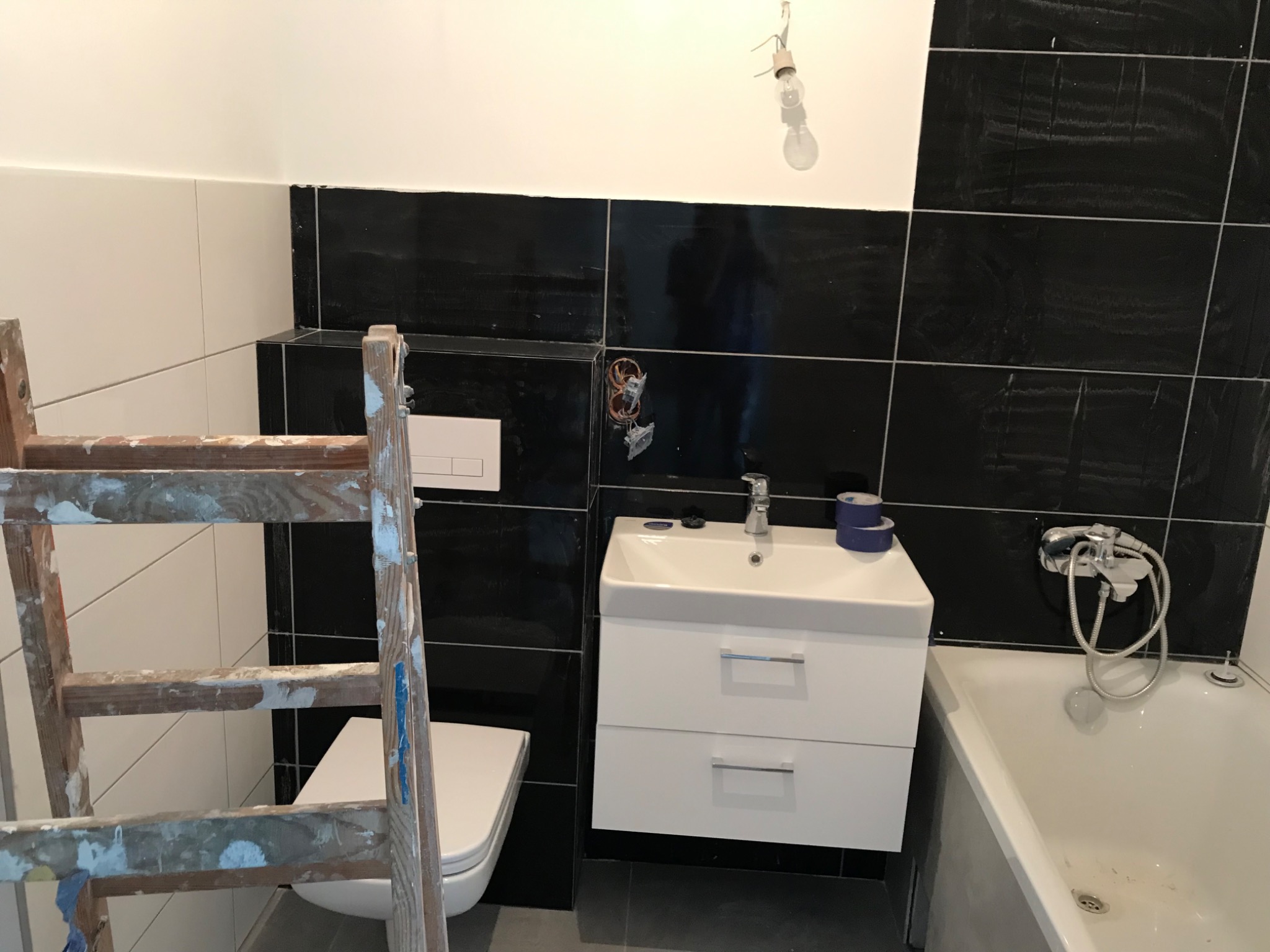 Łazienka w trakcie remontu: widoczna drabina malarska, biała umywalka z szafką, toaleta oraz wanna na tle czarnych płytek, wisząca żarówka.