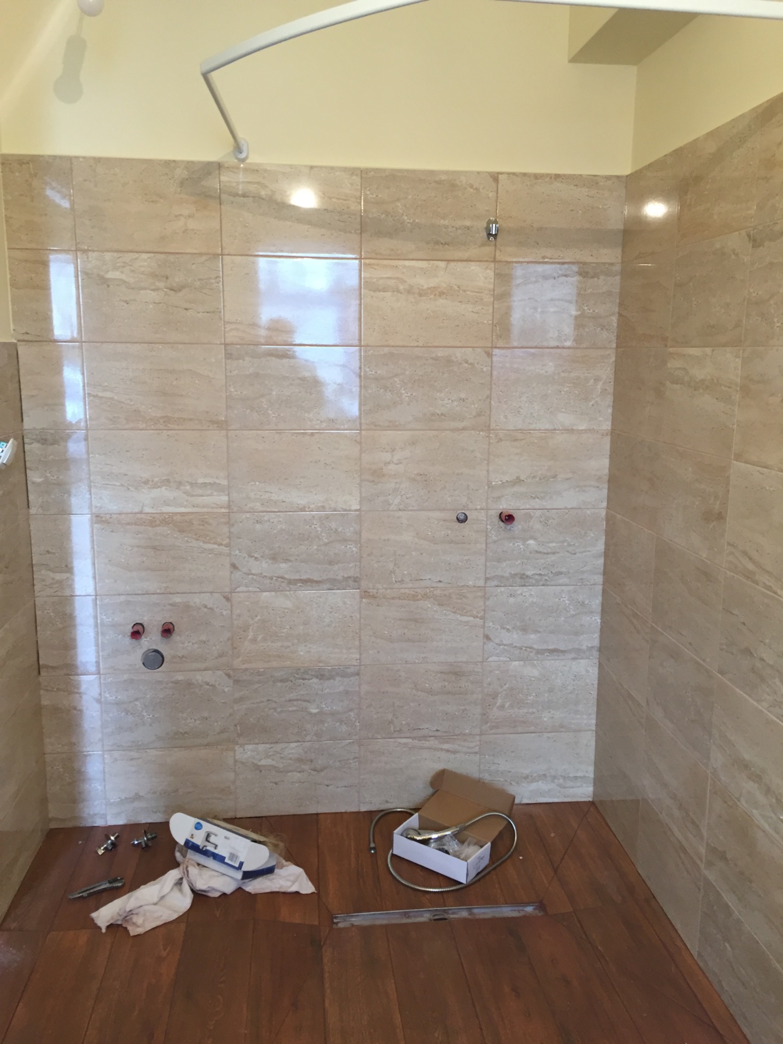 Niedokończona kabina prysznicowa z beżowymi płytkami imitującymi marmur, widoczne przyłącza wodne, biały karnisz, akcesoria do montażu baterii i odpływu na podłodze z paneli imitujących drewno.