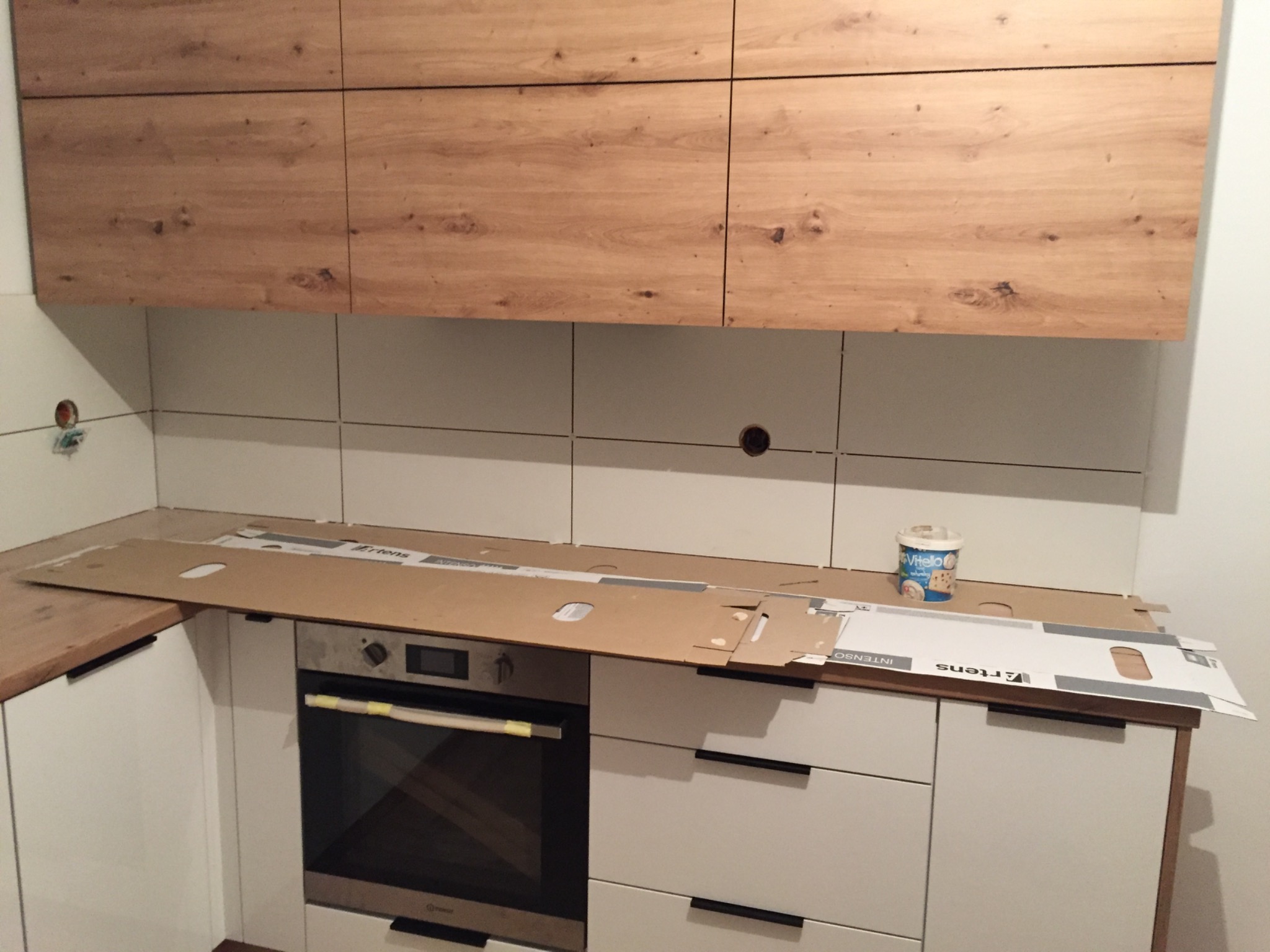 Montaż kuchni: białe szafki z drewnianymi górnymi frontami, zabezpieczony blat z piekarnikiem i puszką po farbie na wierzchu, widoczne płytki na ścianie.