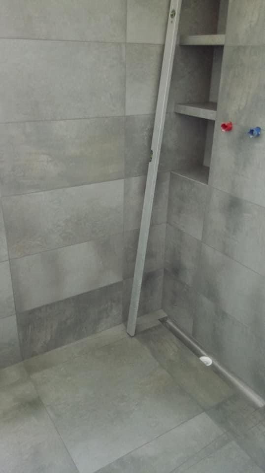 Szara łazienka w trakcie wykańczania, widoczny odpływ liniowy, poziomica oparta o ścianę i wnęka z półkami, przygotowana pod armaturę.