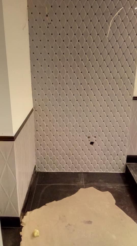Ściana w łazience wyłożona jasnymi, romboidalnymi płytkami z ciemnymi punktami w rogach, widoczny fragment podłogi z ciemnych płytek oraz zabezpieczająca kartonowa mata, w tle widoczny fragment...
