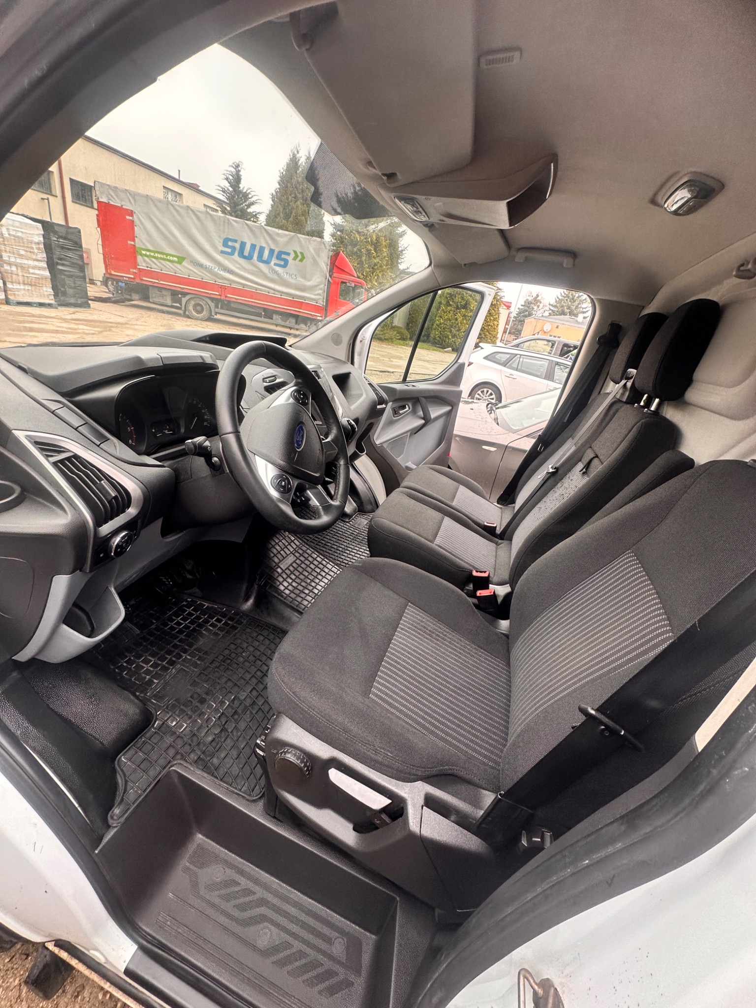 Wnętrze kabiny białego samochodu dostawczego Ford Transit z widocznymi fotelami z szarej tapicerki, gumowymi dywanikami i deską rozdzielczą, z zewnątrz widoczny budynek i ciężarówka SUUS.