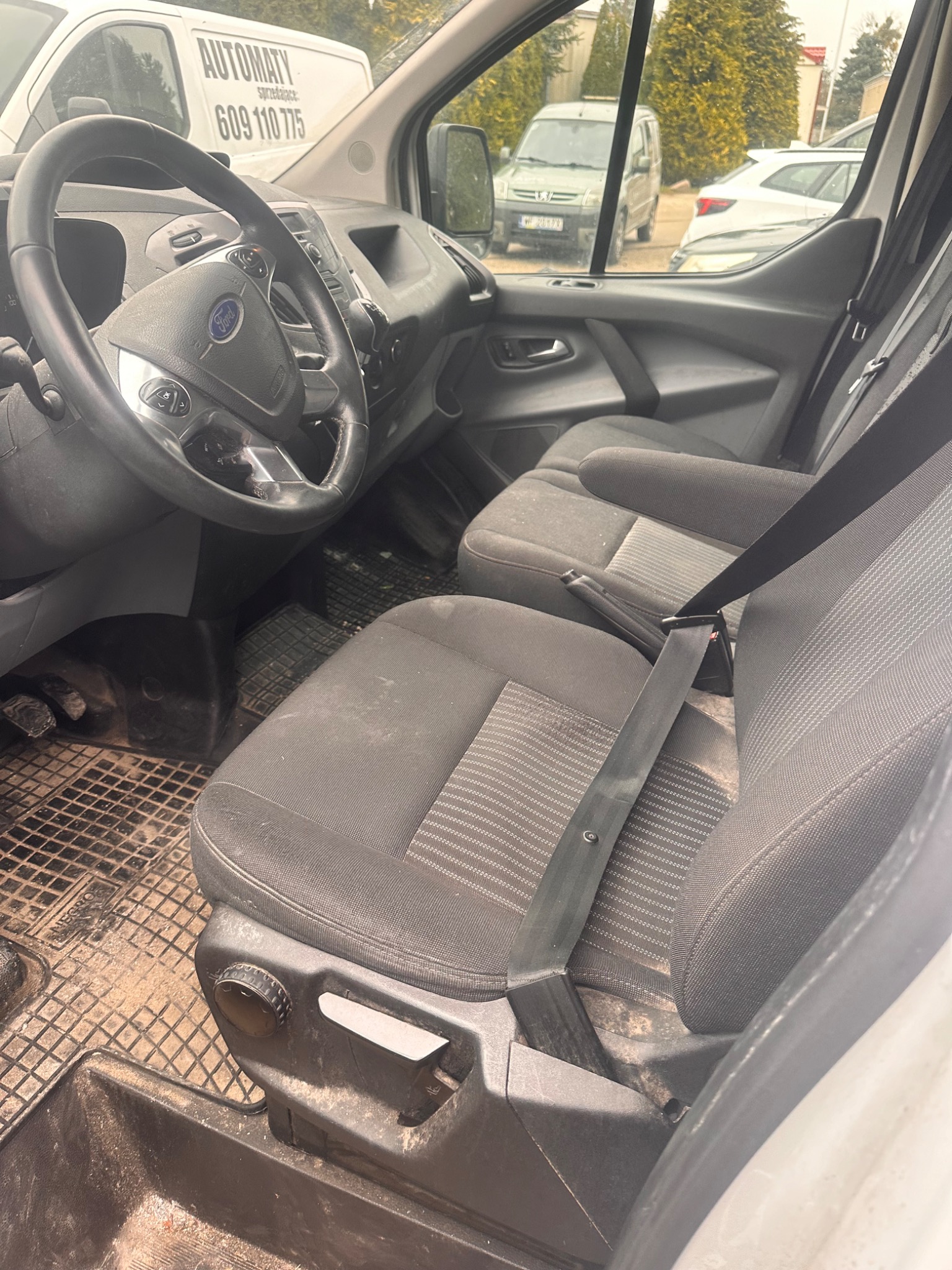 Wnętrze brudnego busa Ford Transit z widocznymi plamami i zabrudzeniami na tapicerce siedzeń oraz podłodze, sugerujące intensywne użytkowanie pojazdu.