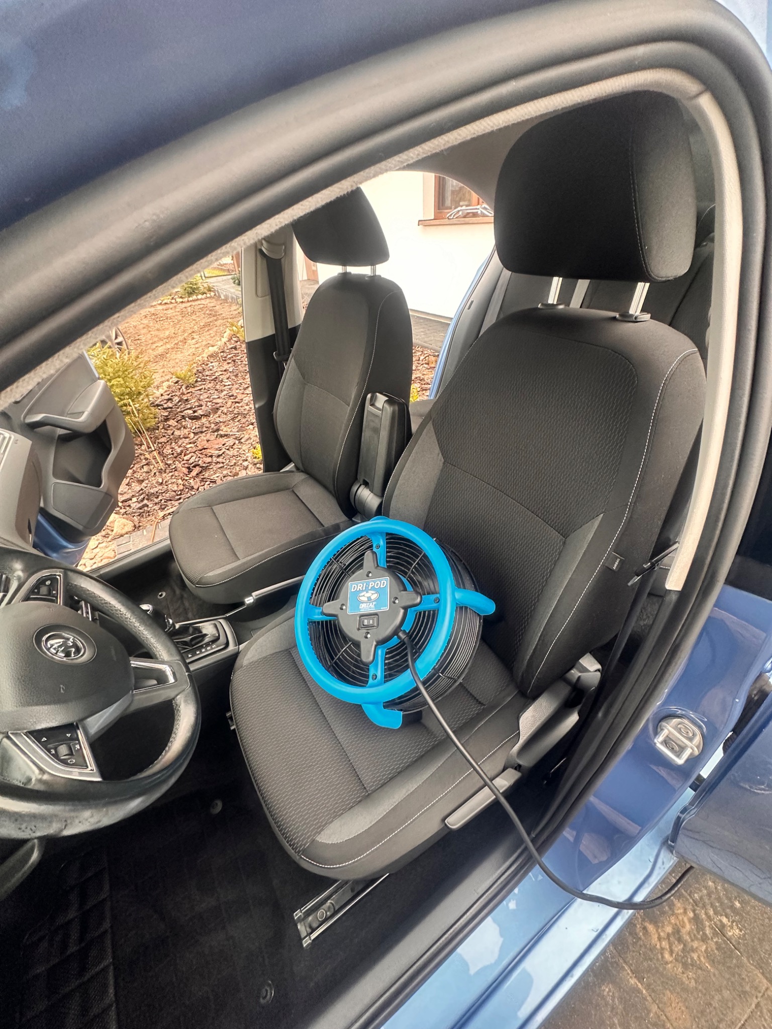 Wnętrze niebieskiego samochodu z czarną tapicerką, na siedzeniu pasażera leży niebieski wentylator marki Dri-Pod podłączony kablem do gniazdka, prawdopodobnie w celu osuszenia po czyszczeniu.