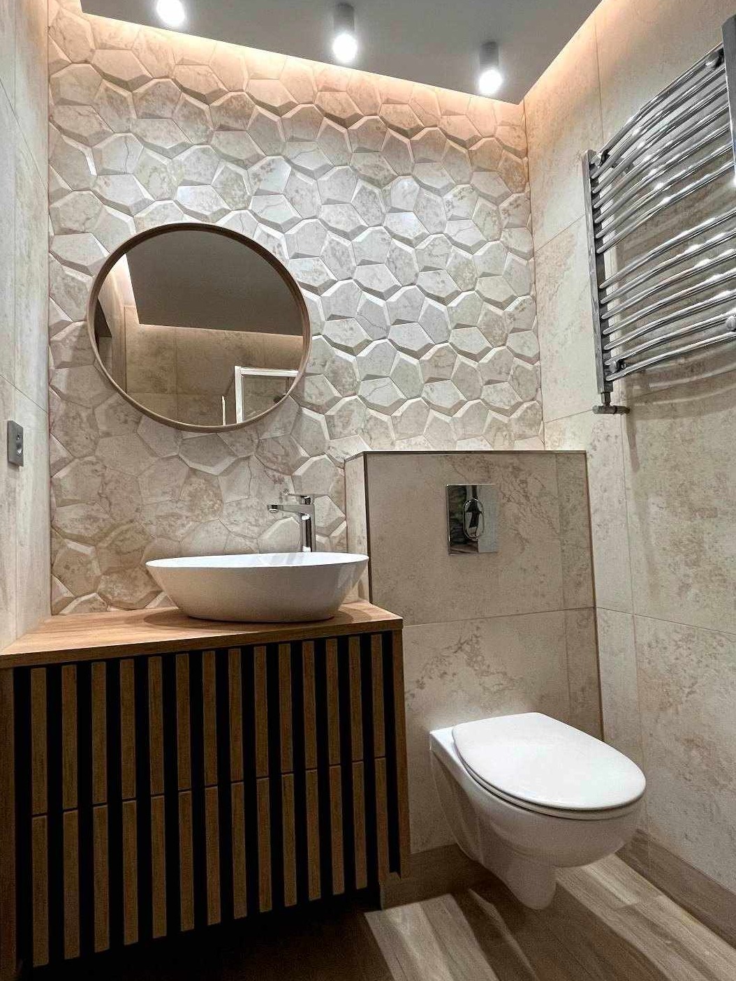 Nowoczesna łazienka z umywalką nablatową na szafce z drewnianymi lamelami, okrągłym lustrem i dekoracyjną mozaiką na ścianie, oświetlona punktowymi lampami sufitowymi.